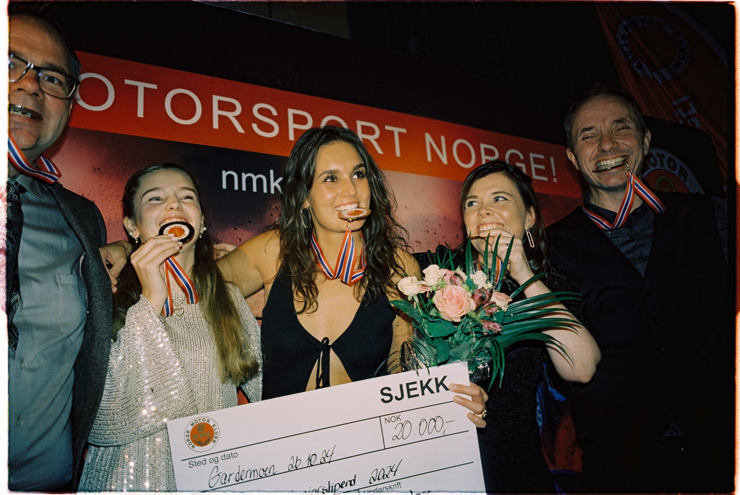 People celebrating a victory, holding medals and a large check, in front of a red backdrop with the text 'MOTORSPOERT NORGE!' and a partial word 'nmk'. The group includes two men and three women, one woman holding a bouquet of flowers, smiling and laughing.