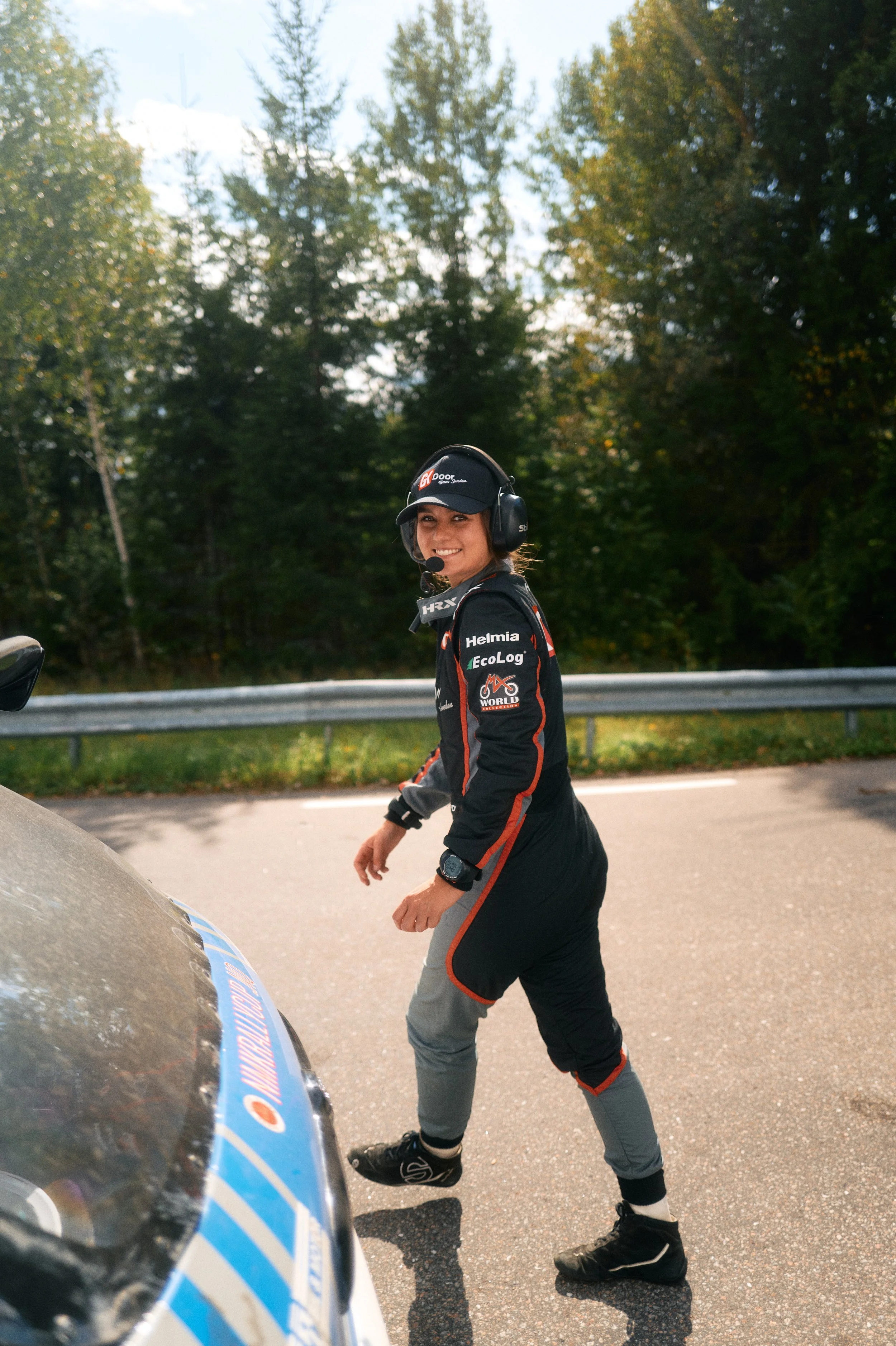A smiling female race car driver wearing a helmet and racing suit, standing next to a race car on a road with trees in the background.