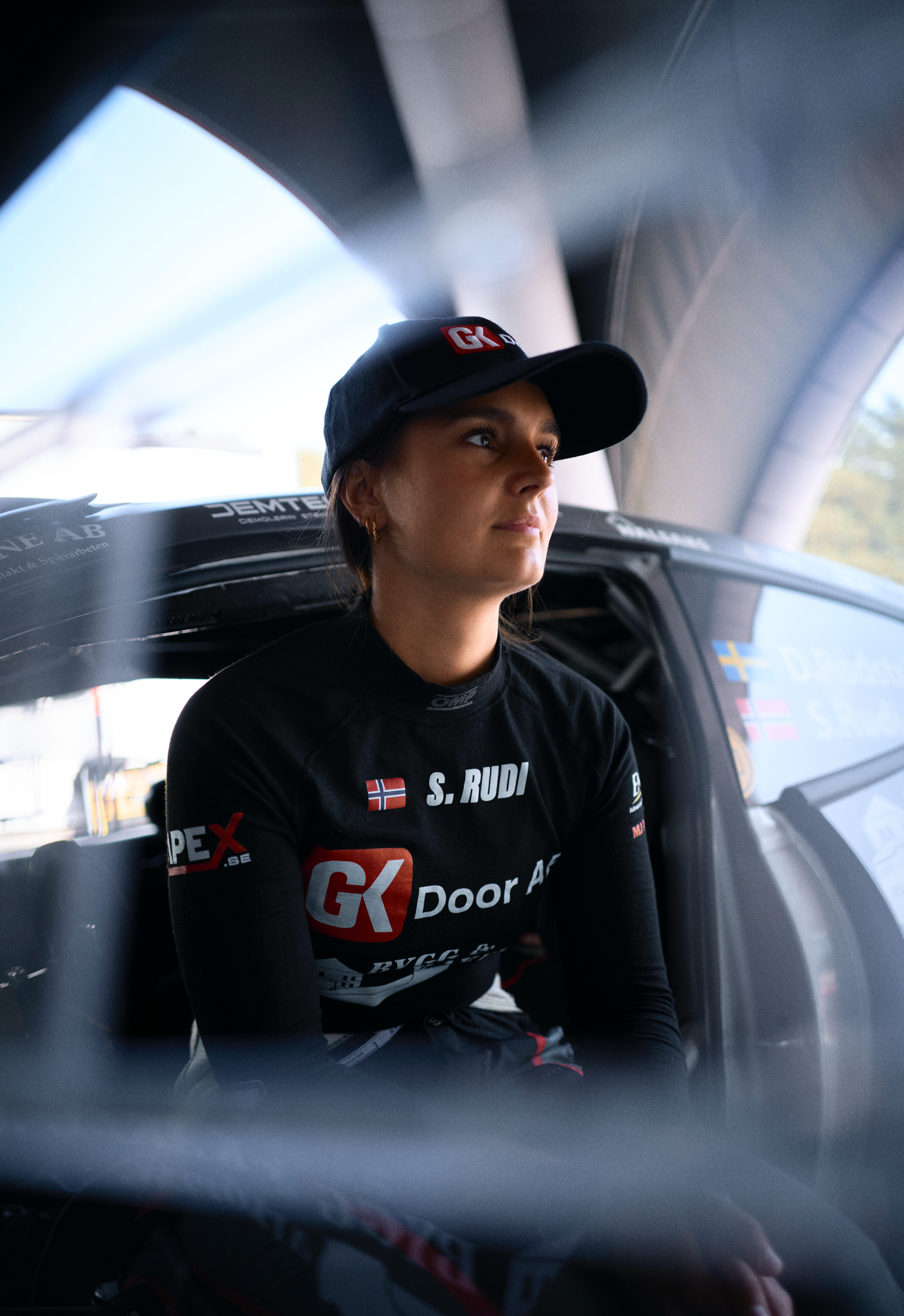 A female race car driver sitting inside a racing vehicle, wearing a black cap, black racing suit with sponsor logos, and looking thoughtfully out of the window.
