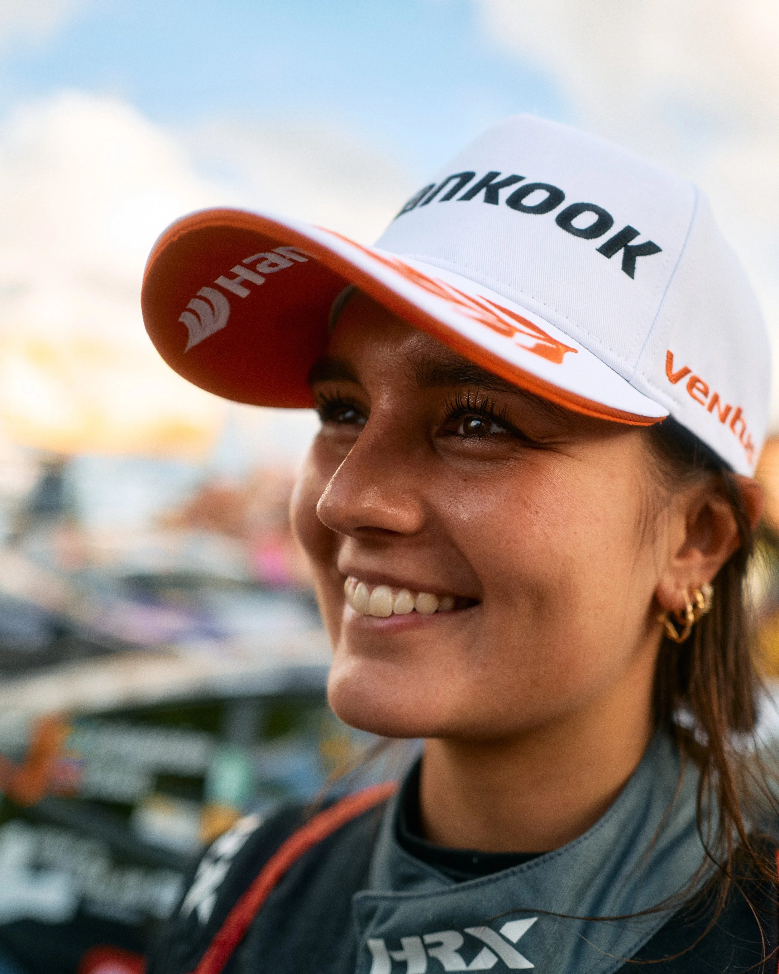 Close-up of a smiling woman wearing a white and orange cap with the logos of HOKA and Venly, outdoors on a sunny day.