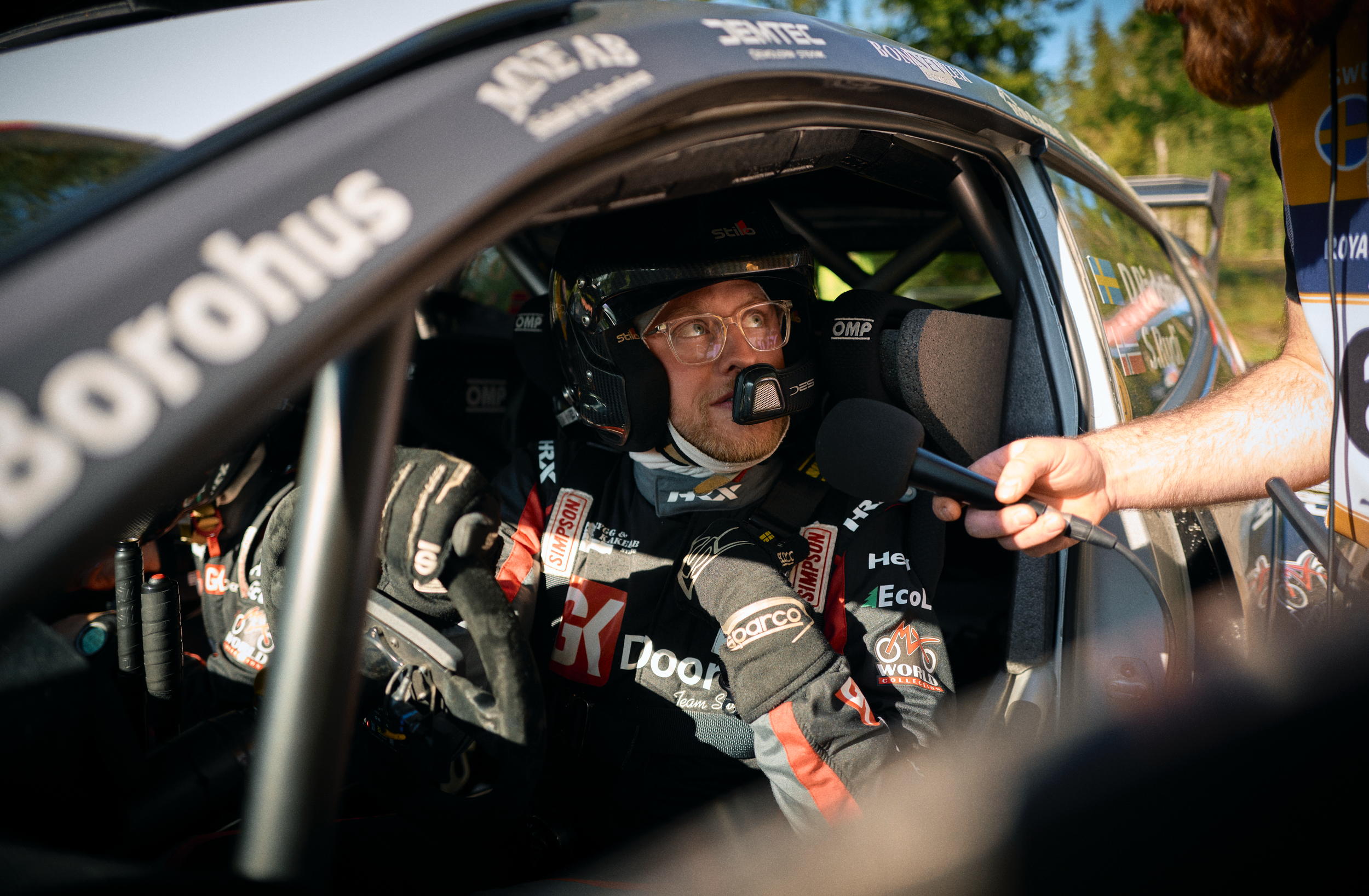 Race car driver in racing suit sitting inside a car, being interviewed with a microphone held close to his face, wearing a helmet and glasses, with race logos and Swedish flag on the car window.