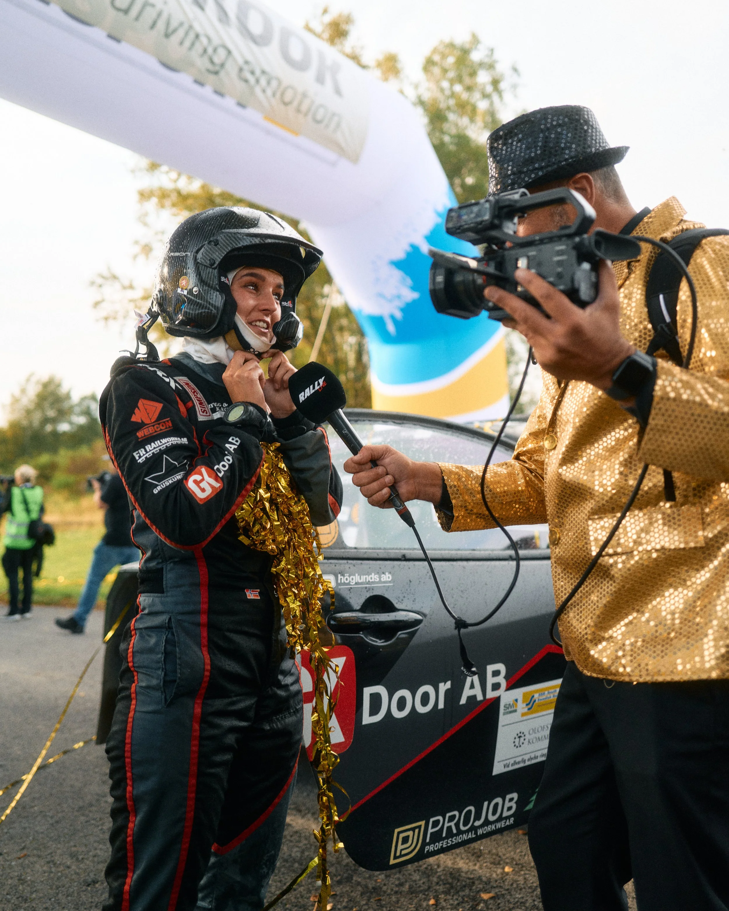 A female rally car driver being interviewed with a microphone, wearing a helmet and a racing suit decorated with gold tinsel, as she celebrates her race.