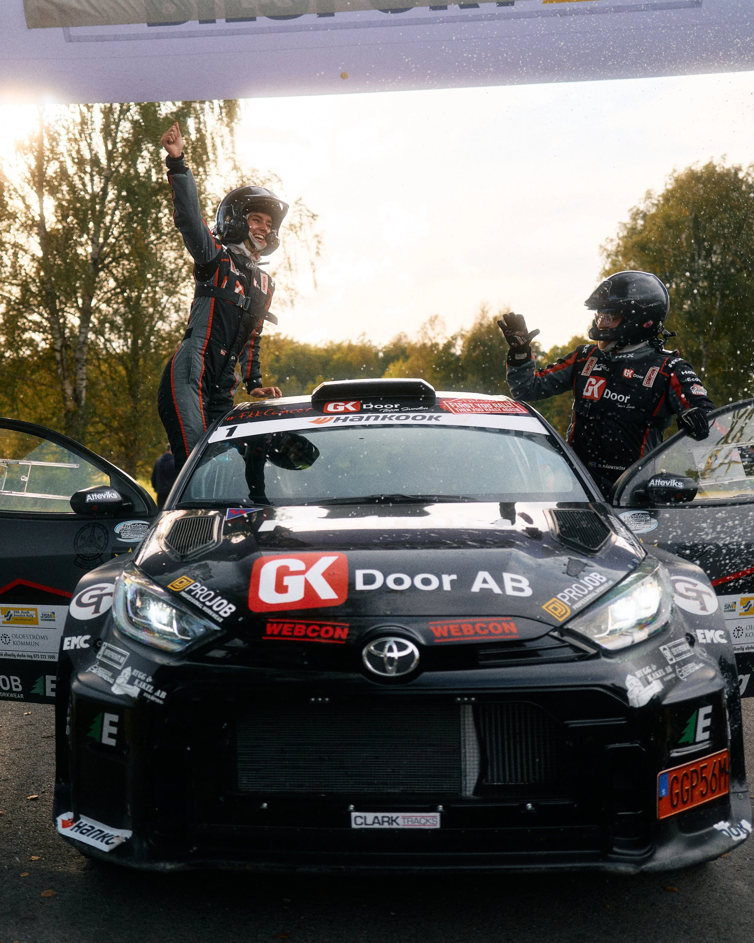 Two race car drivers in racing suits and helmets celebrating on a black Toyota race car after a race, raising their hands and smiling, with trees and autumn sky in the background.