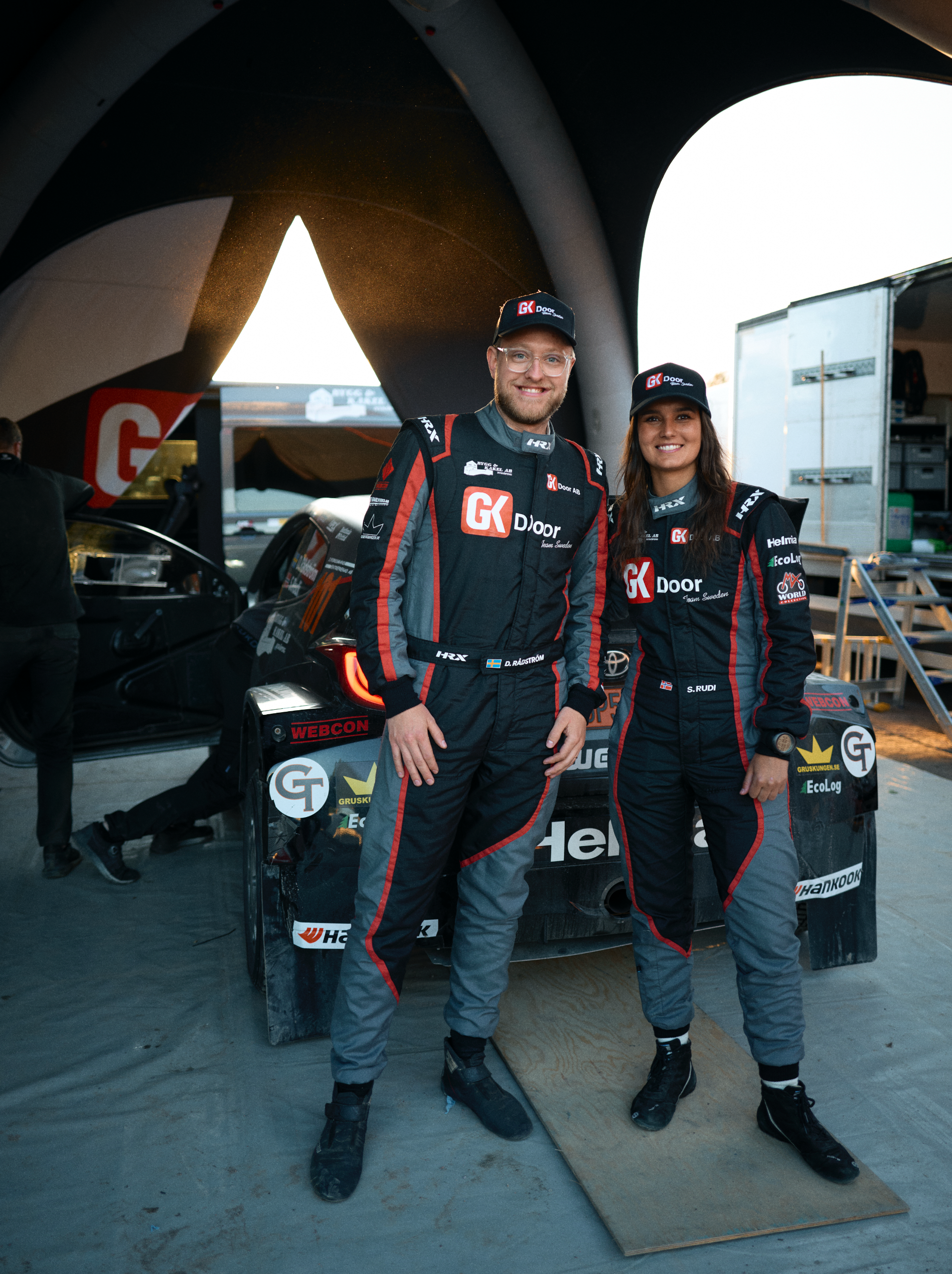 Two race car drivers in racing suits and caps, standing in front of a race car under a large tent at a racing event.