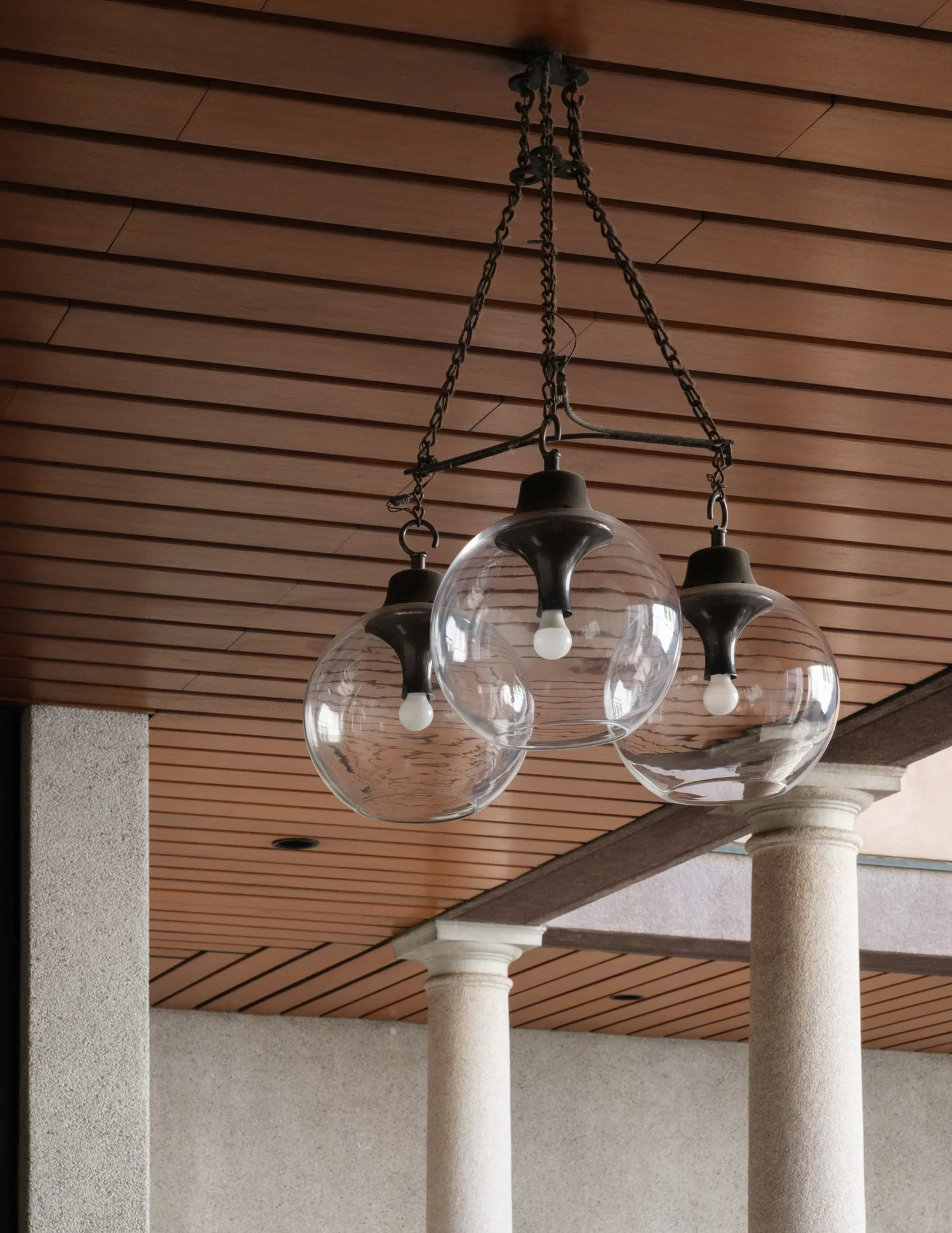 A modern chandelier with three glass globes hanging from a ceiling with wooden slats, supported by two white columns.