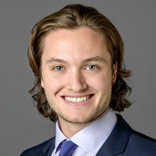 A young man with shoulder-length brown hair, wearing a dark suit, light shirt, and tie, smiling at the camera
