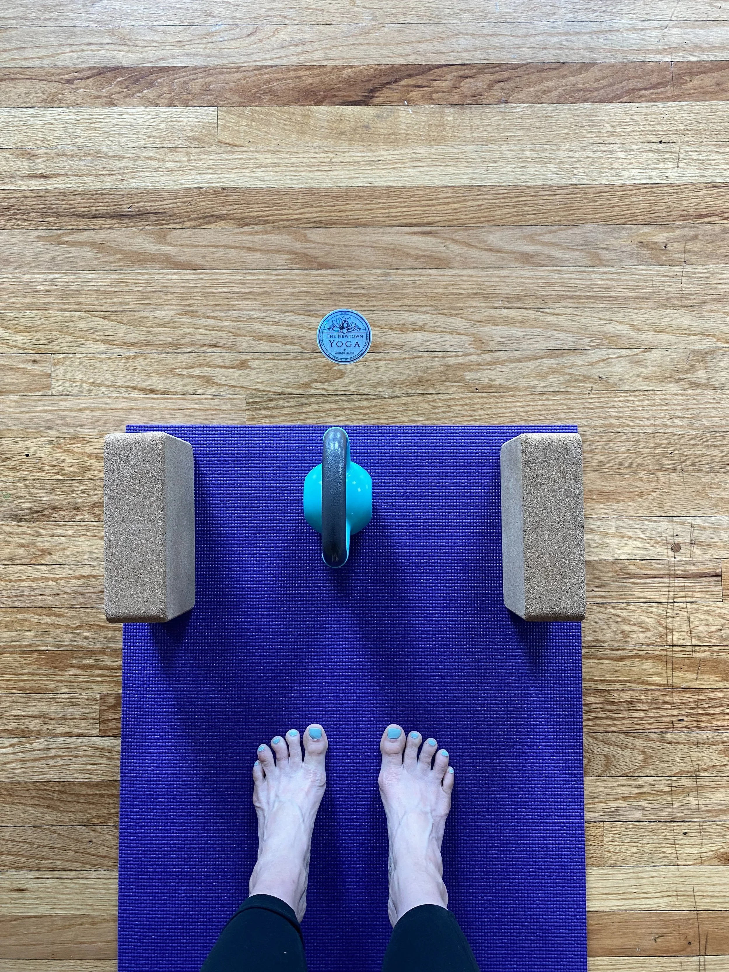 Top-down view of a person standing on a purple yoga mat, feet with painted toenails, with yoga blocks, a kettlebell, and a sticker nearby on a wooden floor.
