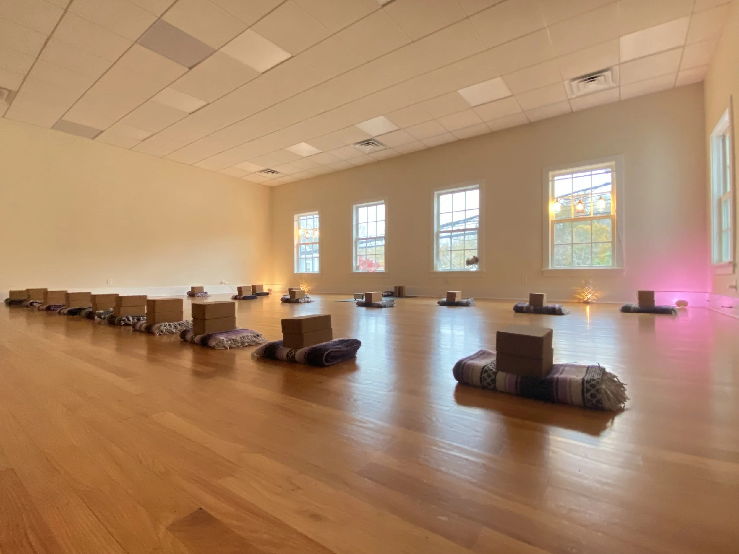 A spacious, well-lit yoga or meditation studio with a polished wooden floor. Yoga mats are rolled out on the floor in neat rows, each topped with a small stack of blocks and a folded blanket. Large windows along the wall let in natural light, and small decorative lights are on the floor near the walls.