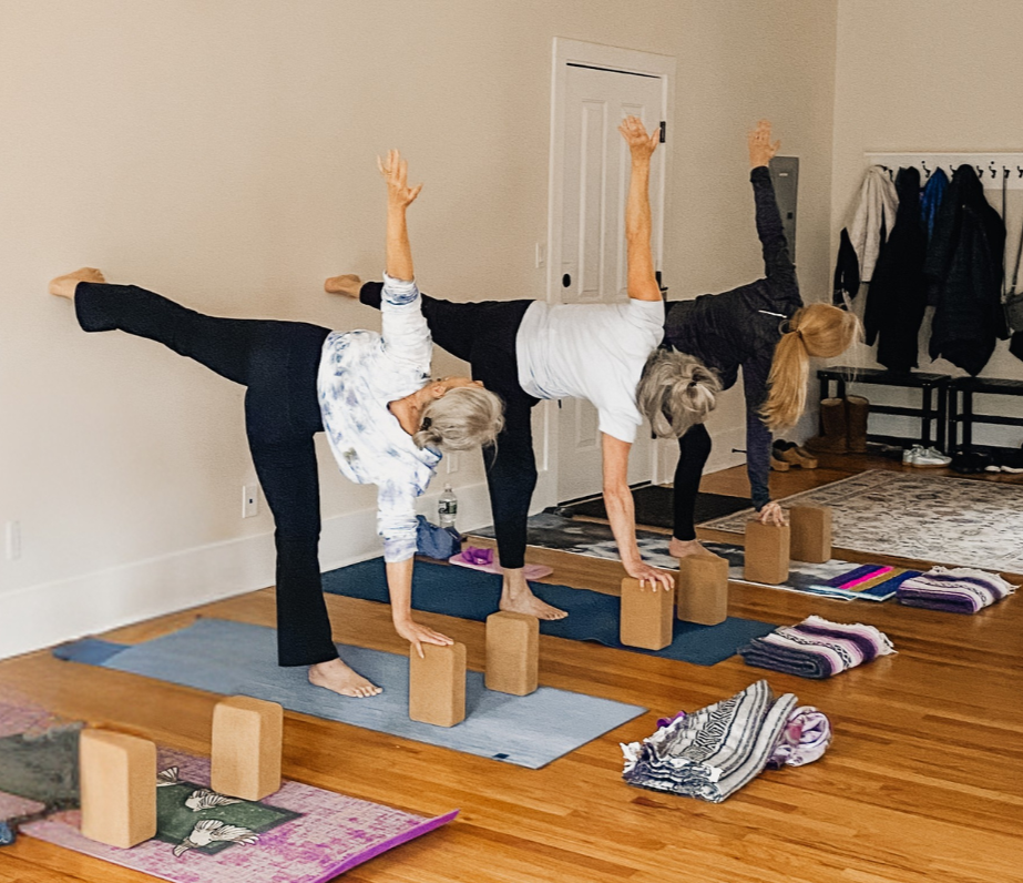 Three women practicing yoga on mats, balancing on one leg while holding their other leg and stretching upward, using yoga blocks for support in a spacious living room.