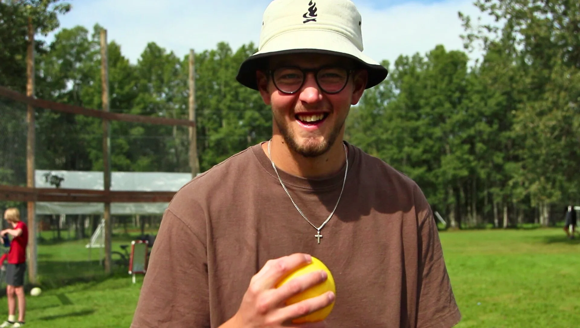Lächelnder junger Mann mit Brille, Hut und Ketten mit Kreuz, hält einen gelben Ball, im Freien auf grünem Rasen bei sonnigem Wetter.