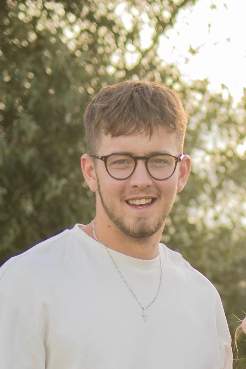 Ein junger Mann mit Brille, kurzer brauner Hair, weißen T-Shirt und Kreuzkette lächelnd im Freien bei Sonnenuntergang.