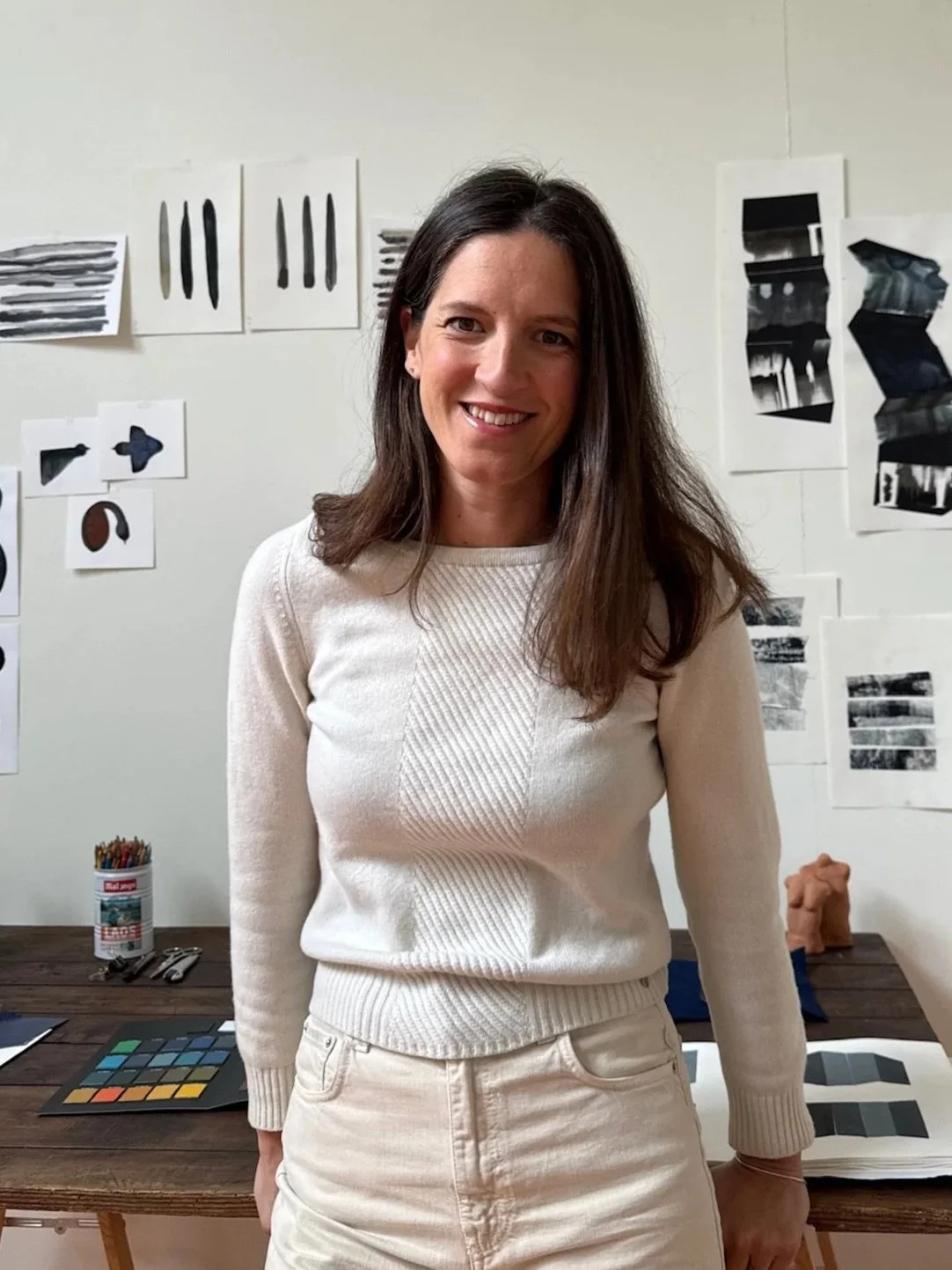 Une femme souriante avec des cheveux bruns portait un pull blanc, se tenant dans un studio avec des recherches visuelles en noir et blanc sur les murs.