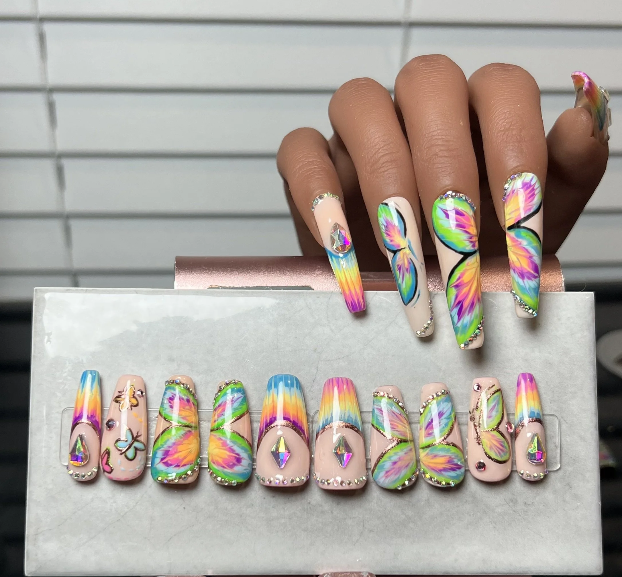 Hand with rainbow-colored butterfly nail art, holding a box of matching butterfly nail designs, featuring vibrant colors and rhinestone accents.