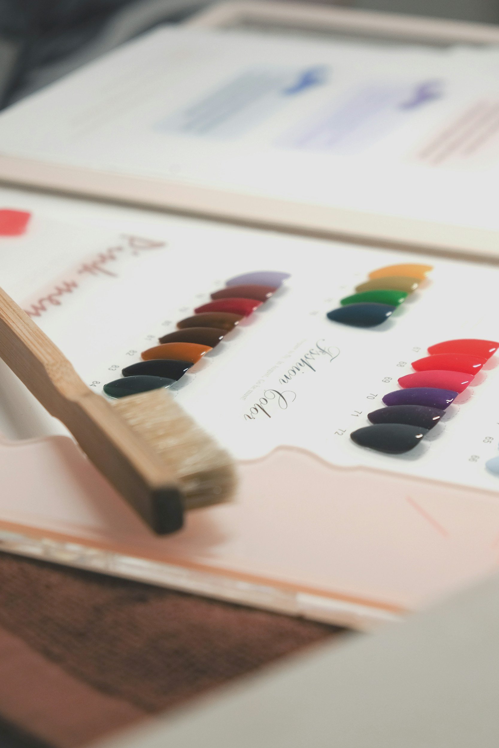 Close-up of a nail polish color palette with various shades of red, purple, green, yellow, and black, arranged in rows on a white background. Part of a color guide or swatch book is visible in the background.