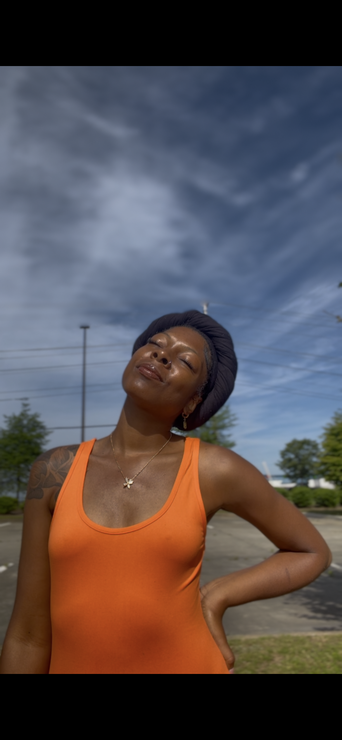 A woman wearing an orange tank top and black headwrap is standing outdoors, smiling with her eyes closed against a cloudy sky backdrop.