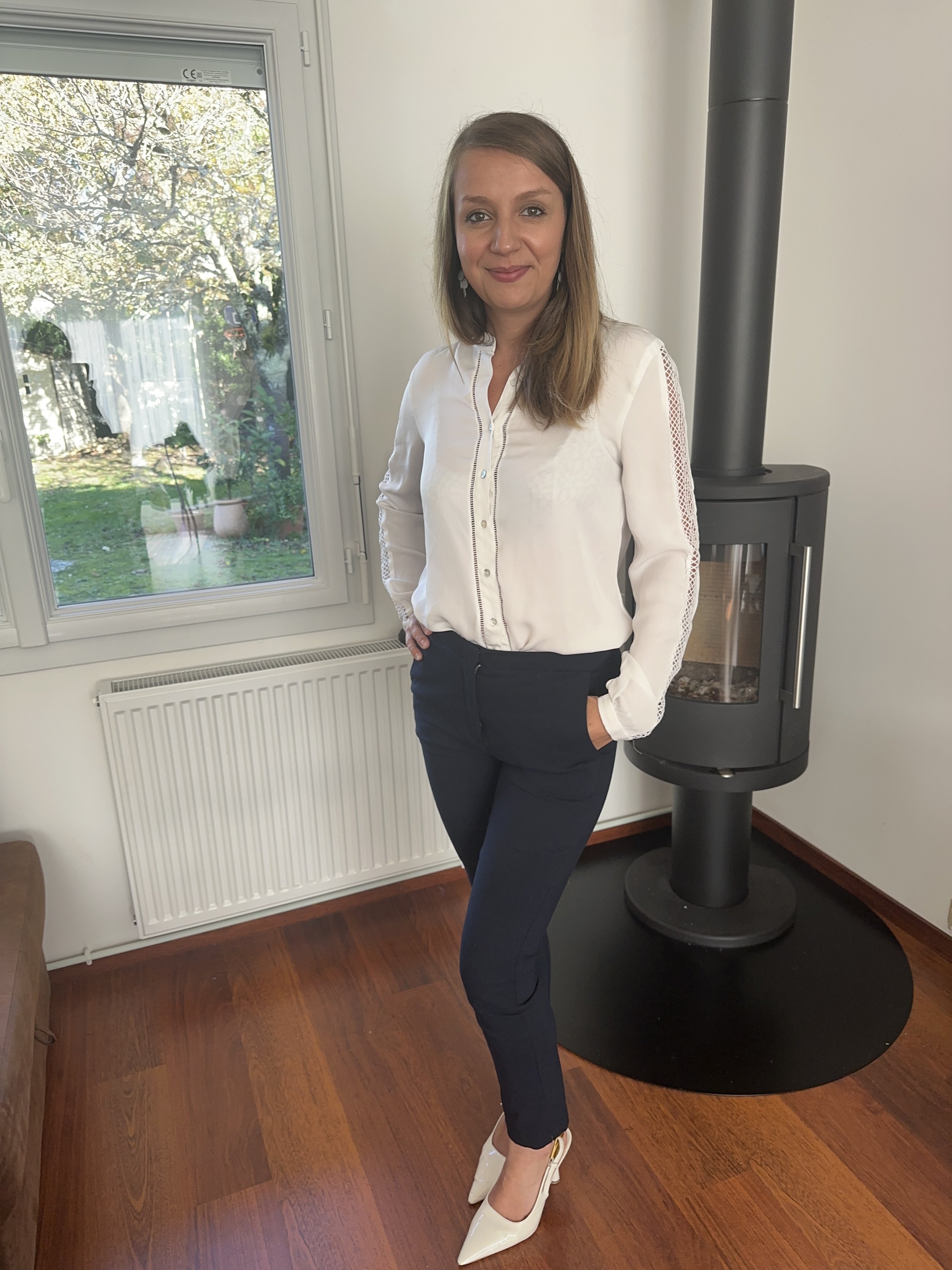 Femme debout à côté d'un poêle à bois dans une pièce, portant une chemise blanche et un pantalon noir, avec des talons blancs.