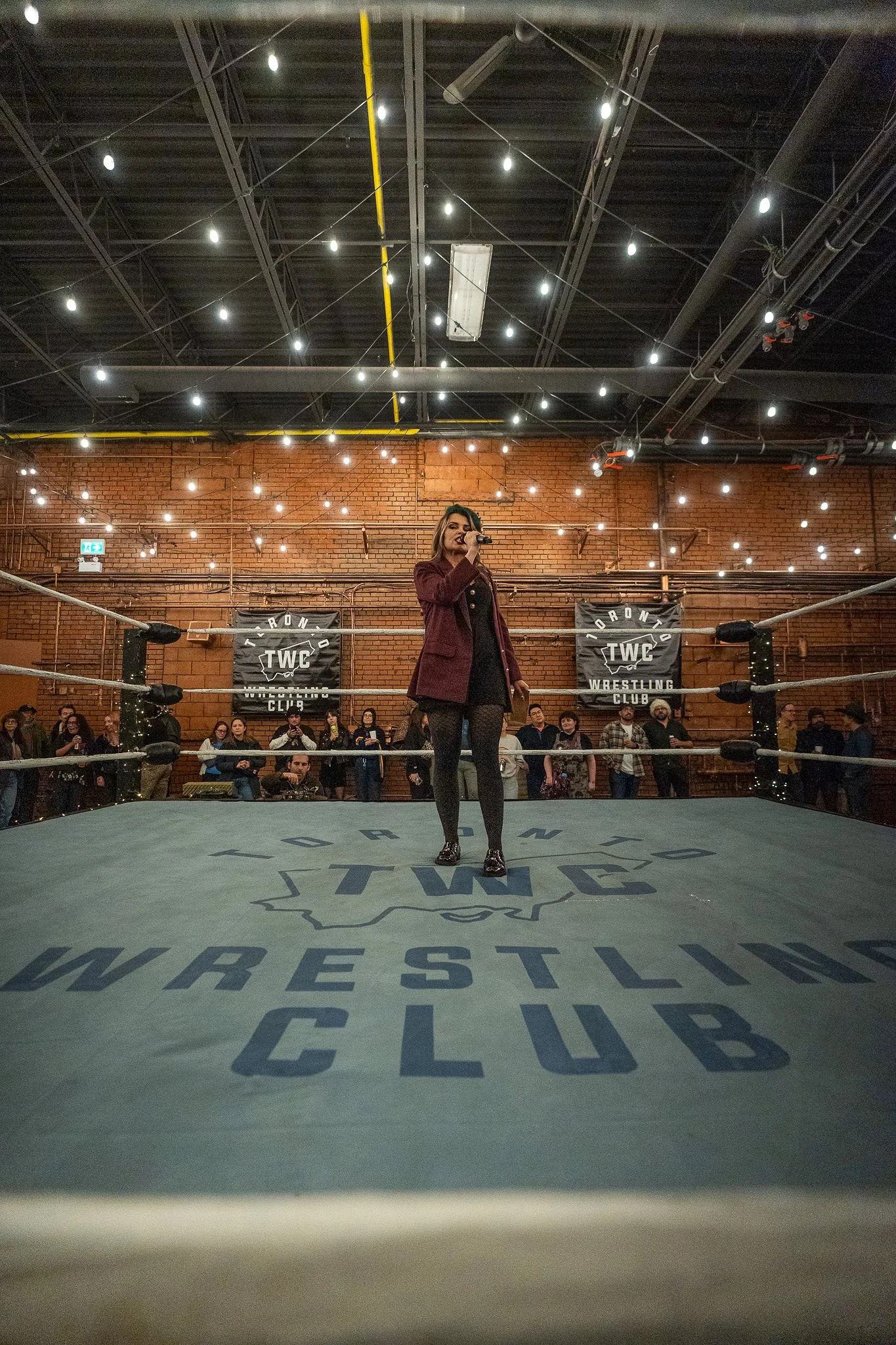 A woman in a wrestling ring speaking into a microphone at Toronto Wrestling Club event, with spectators in the background and lights overhead.