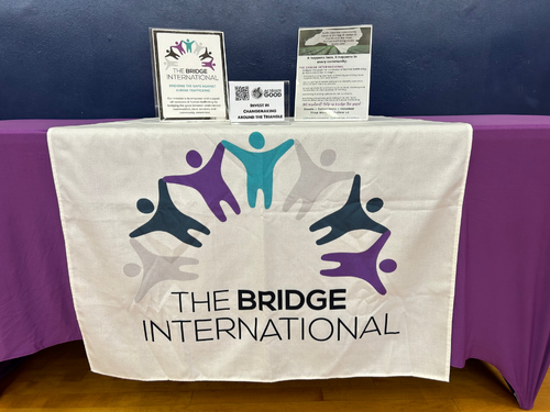 Table with a white cloth featuring the Bridge International logo with colorful abstract figures. On the table are informational pamphlets and signs, with a purple tablecloth in the background.