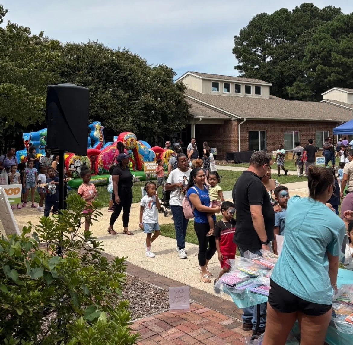 Crowd of people, including children and adults, at an outdoor event with inflatable rides, tables with items, and a building in the background.