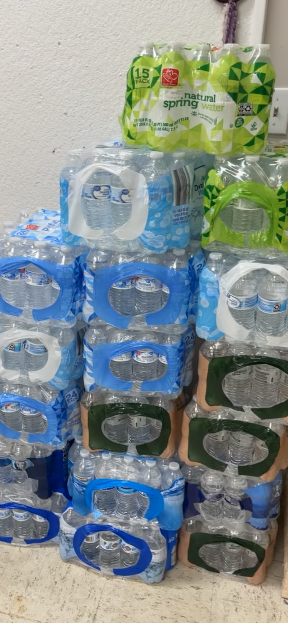 Stacks of bottled water in plastic packaging, with brass-colored cartons mixed in, placed against a wall.