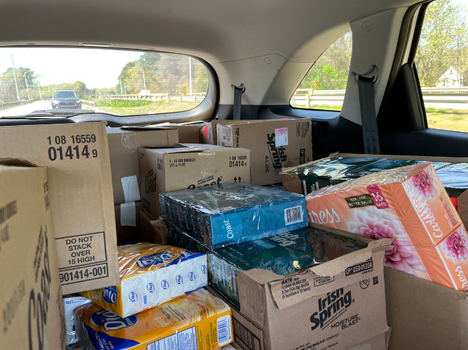 Backseat of a car filled with boxes of household items, including paper towels, tissues, and cleaning supplies, during daytime.