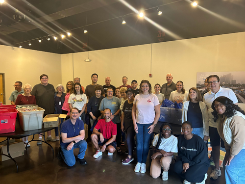 Group of diverse people gathered in a room with hanging lights, some seated on the floor and some standing, in a social or community event setting.