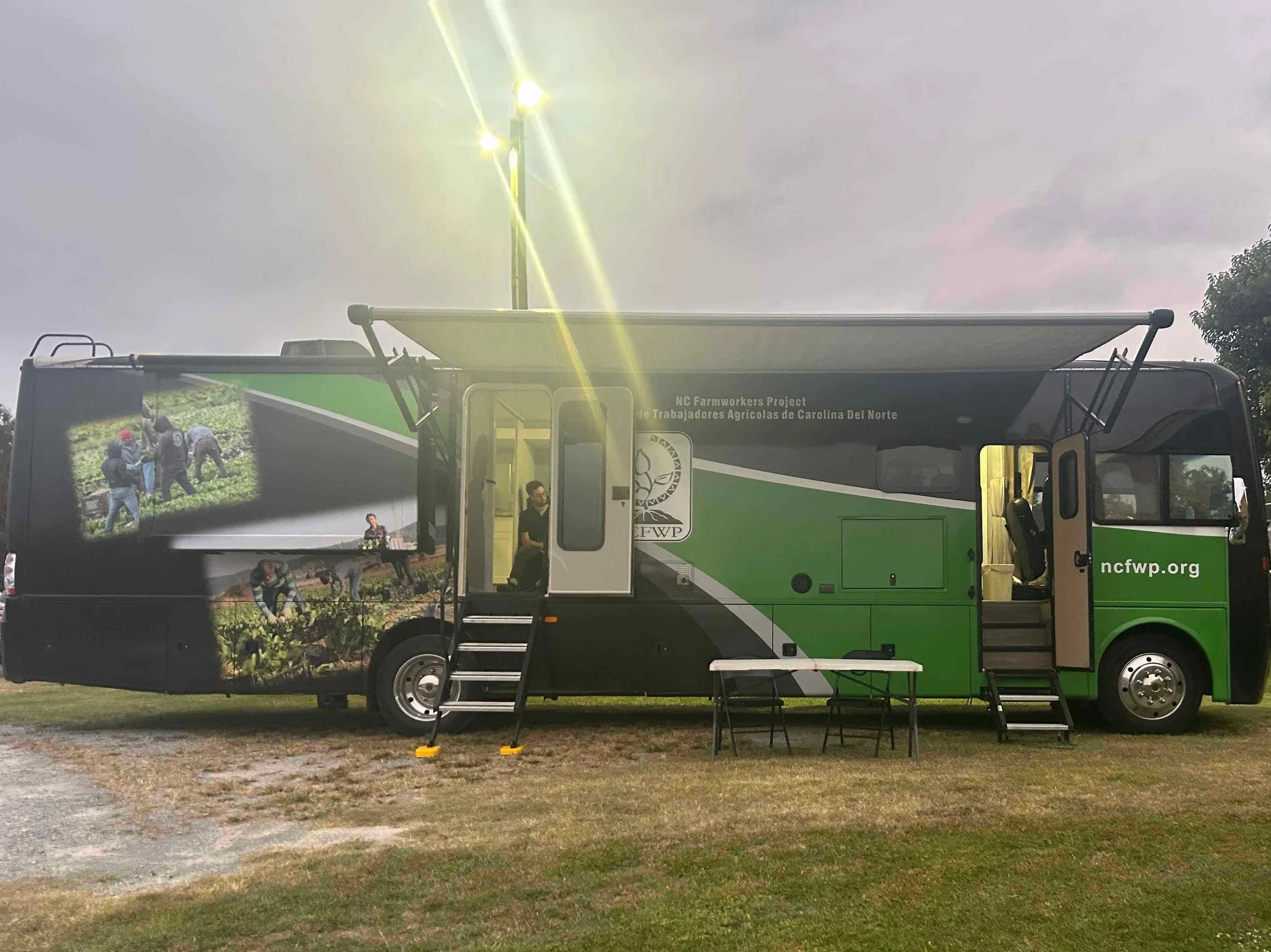 A green and black mobile unit with the logo of NC Farmworkers Project and people working in fields depicted on the side, with an open door, small steps, tables outside, and a daytime outdoor setting under cloudy skies.