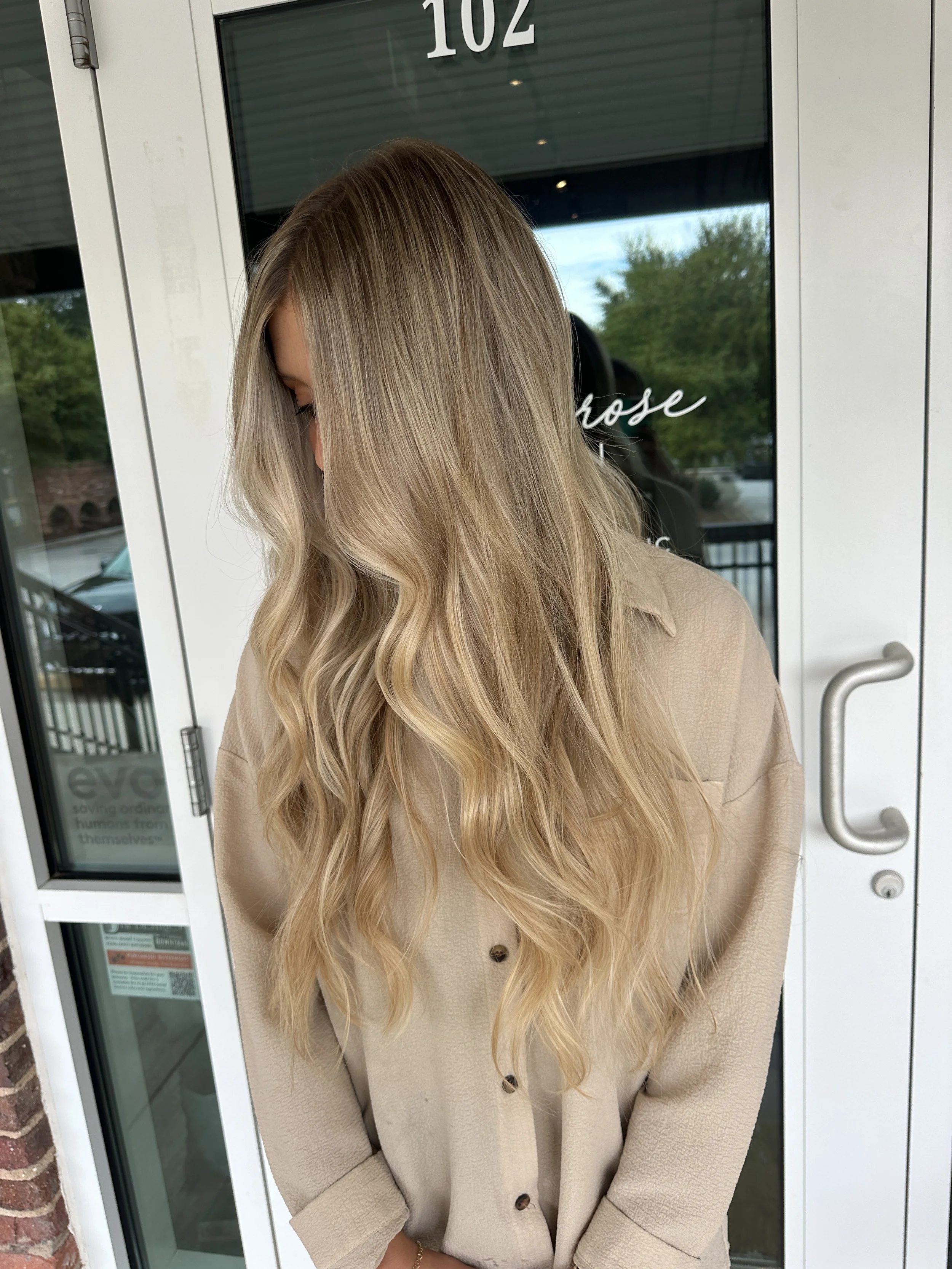 A woman with long, wavy blonde hair standing outdoors in front of a glass door, wearing a beige button-up shirt.