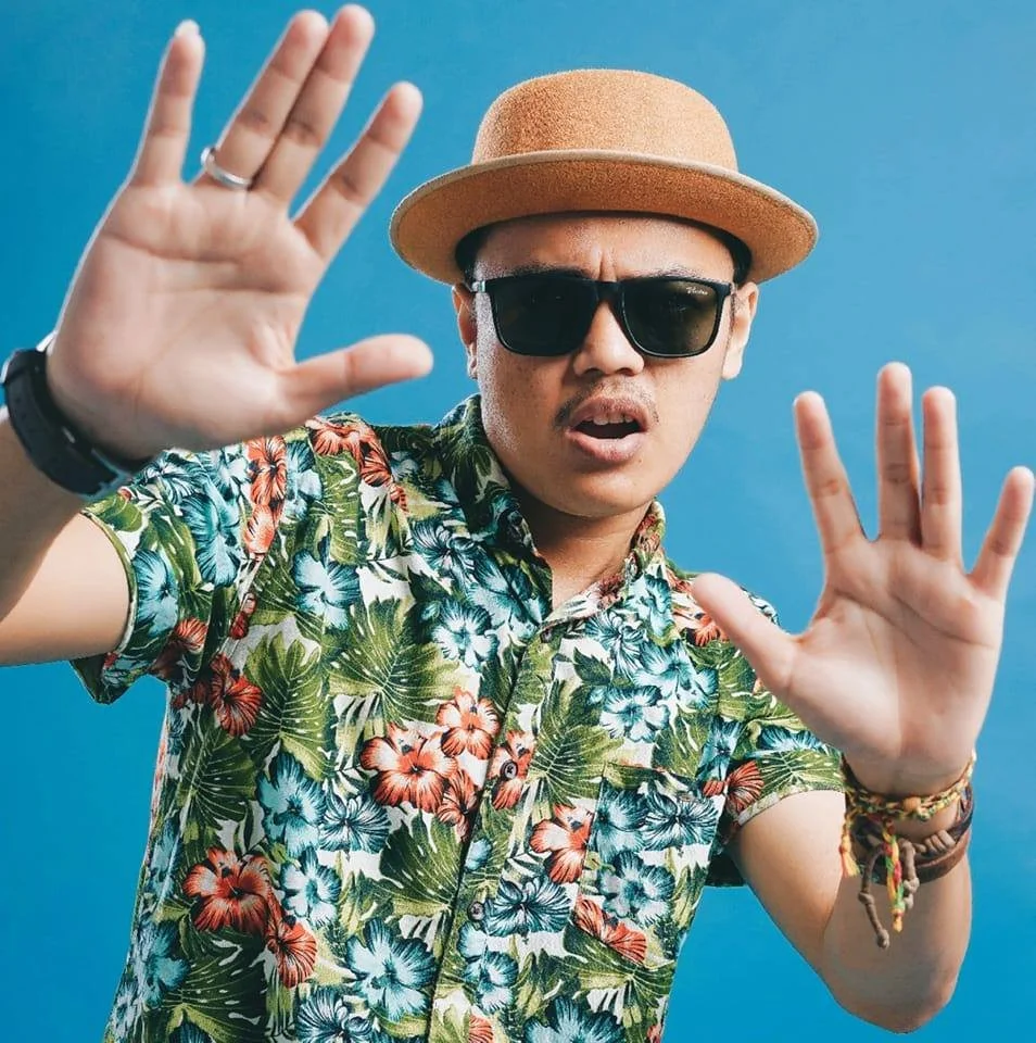 Young man wearing sunglasses, a straw hat, and a tropical floral shirt, raising his hands with a surprised expression against a blue background.