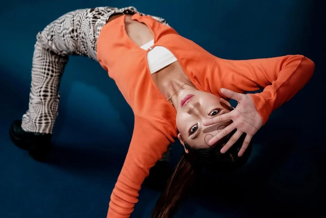 A woman with long dark hair lying on her side on a blue surface, wearing an orange jacket, patterned pants, and a white top, with her hand partially covering her face.