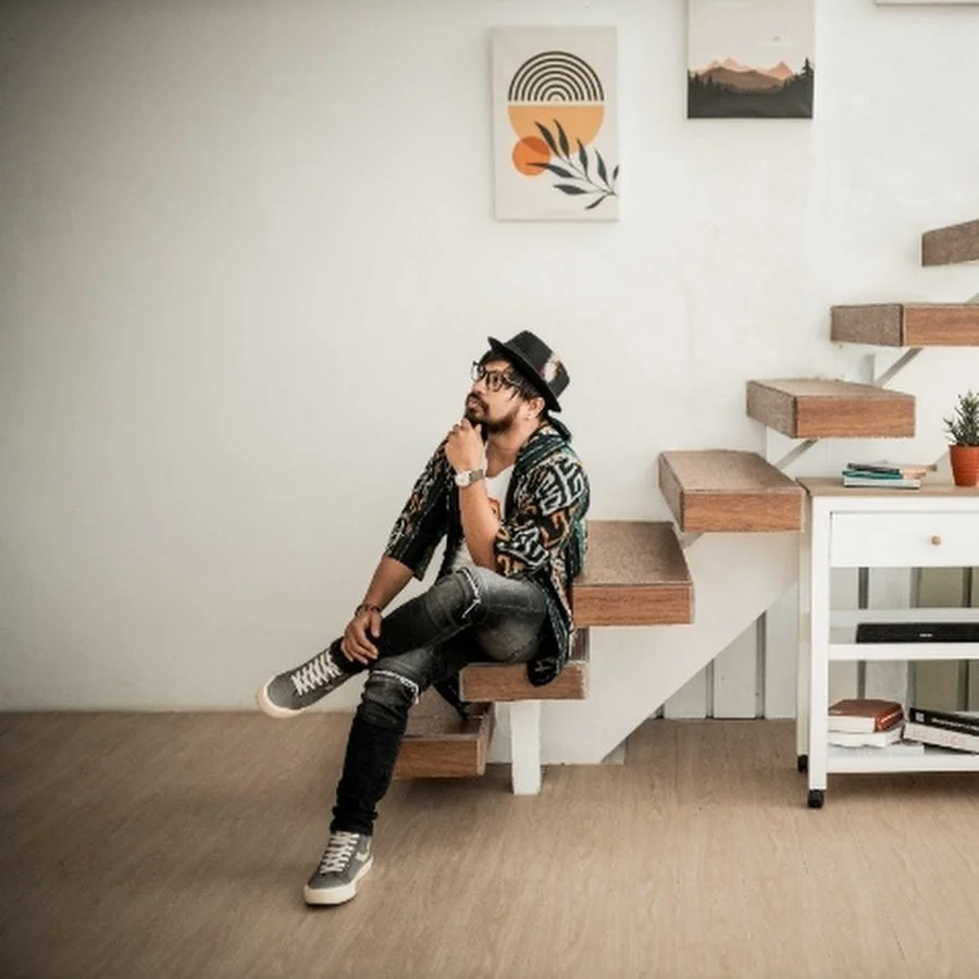 A young man sitting on a set of wooden stairs in a modern, minimalist room. He's wearing glasses, a fedora hat, a patterned cardigan, ripped jeans, and sneakers. The room has a white wall with abstract art and a small white table with books and a potted plant.