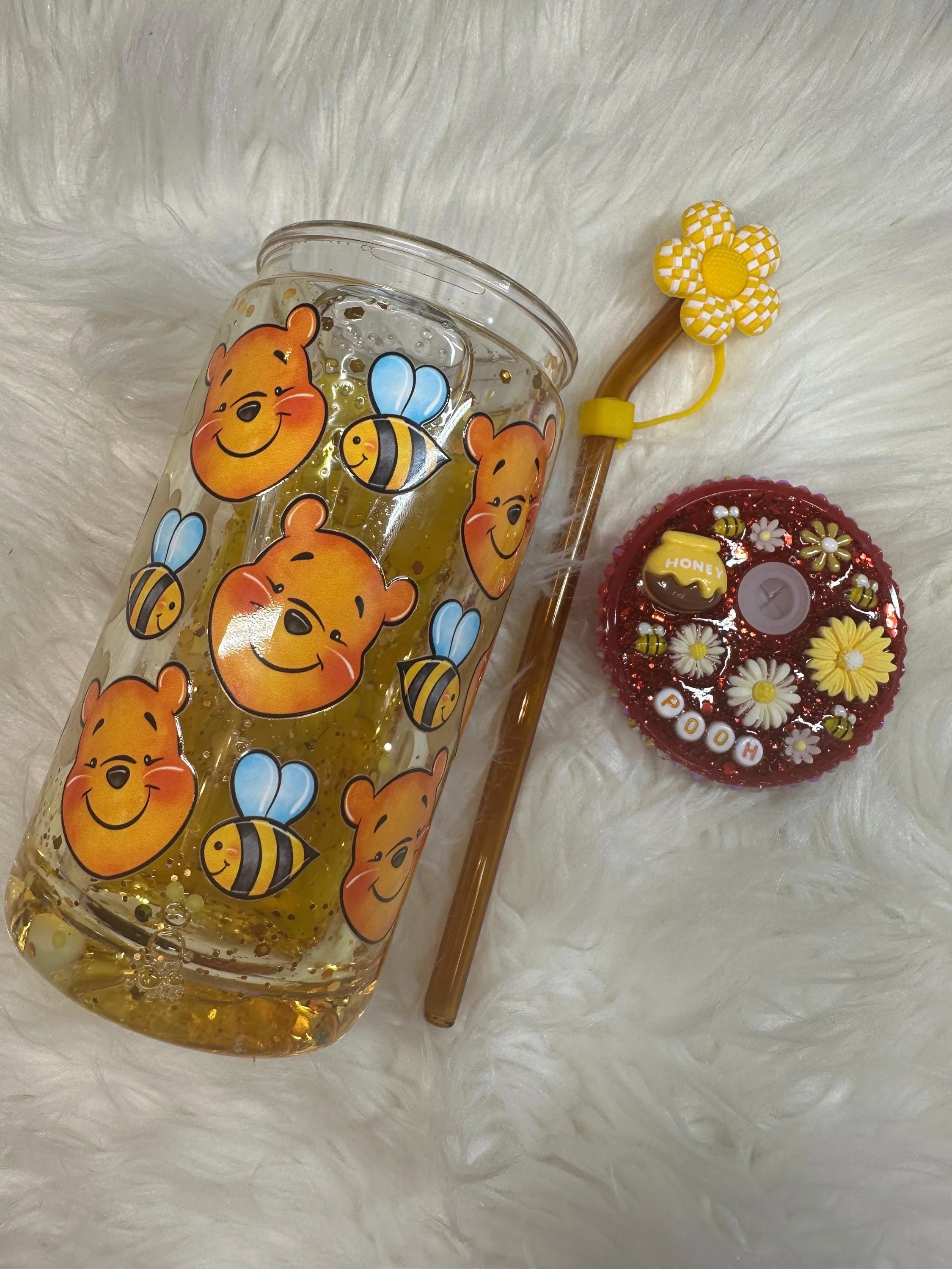 A decorated glass filled with honey, featuring Winnie the Pooh and bees stickers. Next to the glass is a honey dipper with a yellow flower-shaped top and a yellow string. There is also a small round red container with a toy honey jar, flowers, and the words 'POOH' and 'HONEY' on it, placed on a white, fluffy surface.
