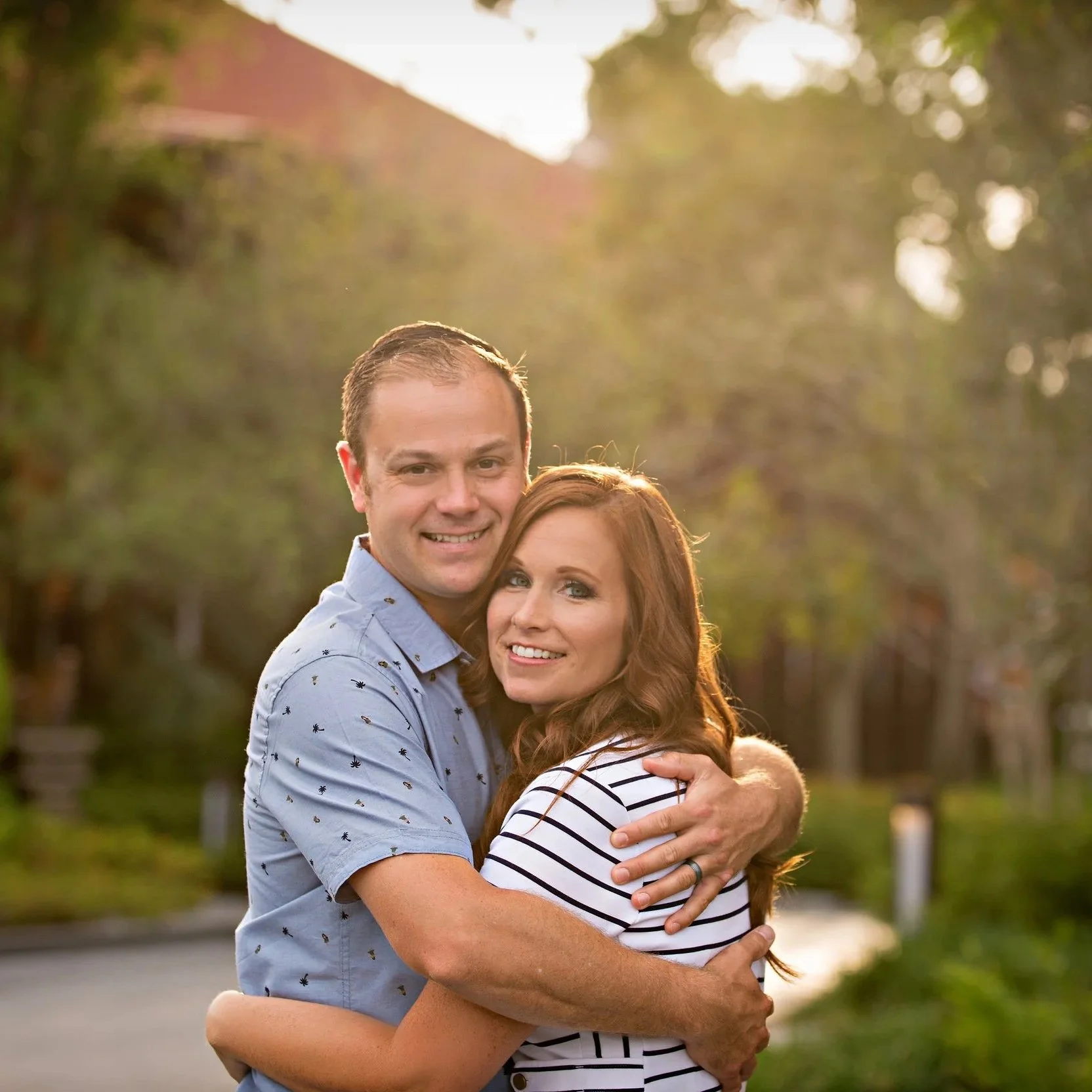 A couple hugging outdoors during sunset, with trees and a road in the background.