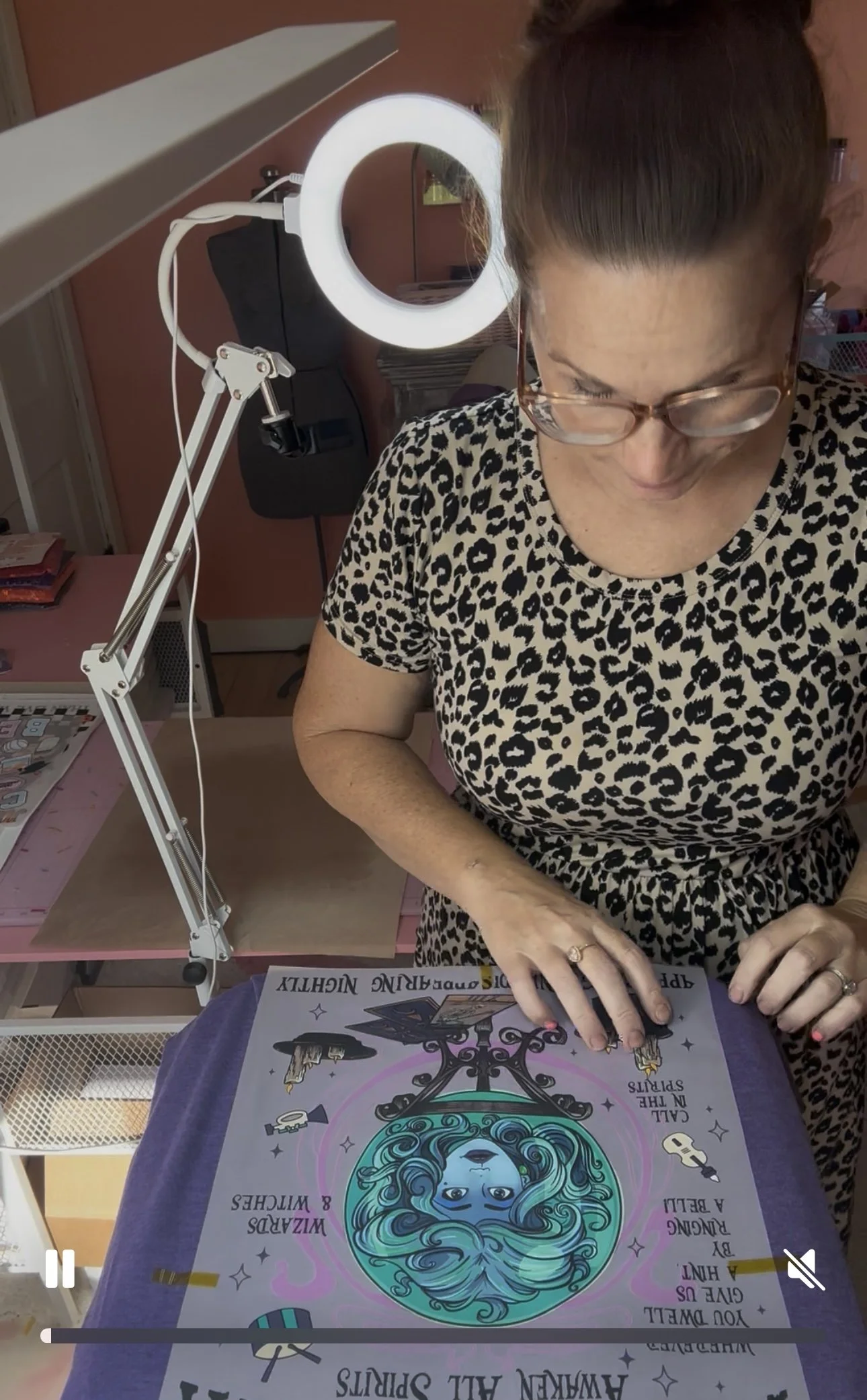 A woman with glasses and a leopard print shirt is working on a colorful illustration of a woman with teal hair and a mirror, surrounded by text and decorative elements, under a ring light.