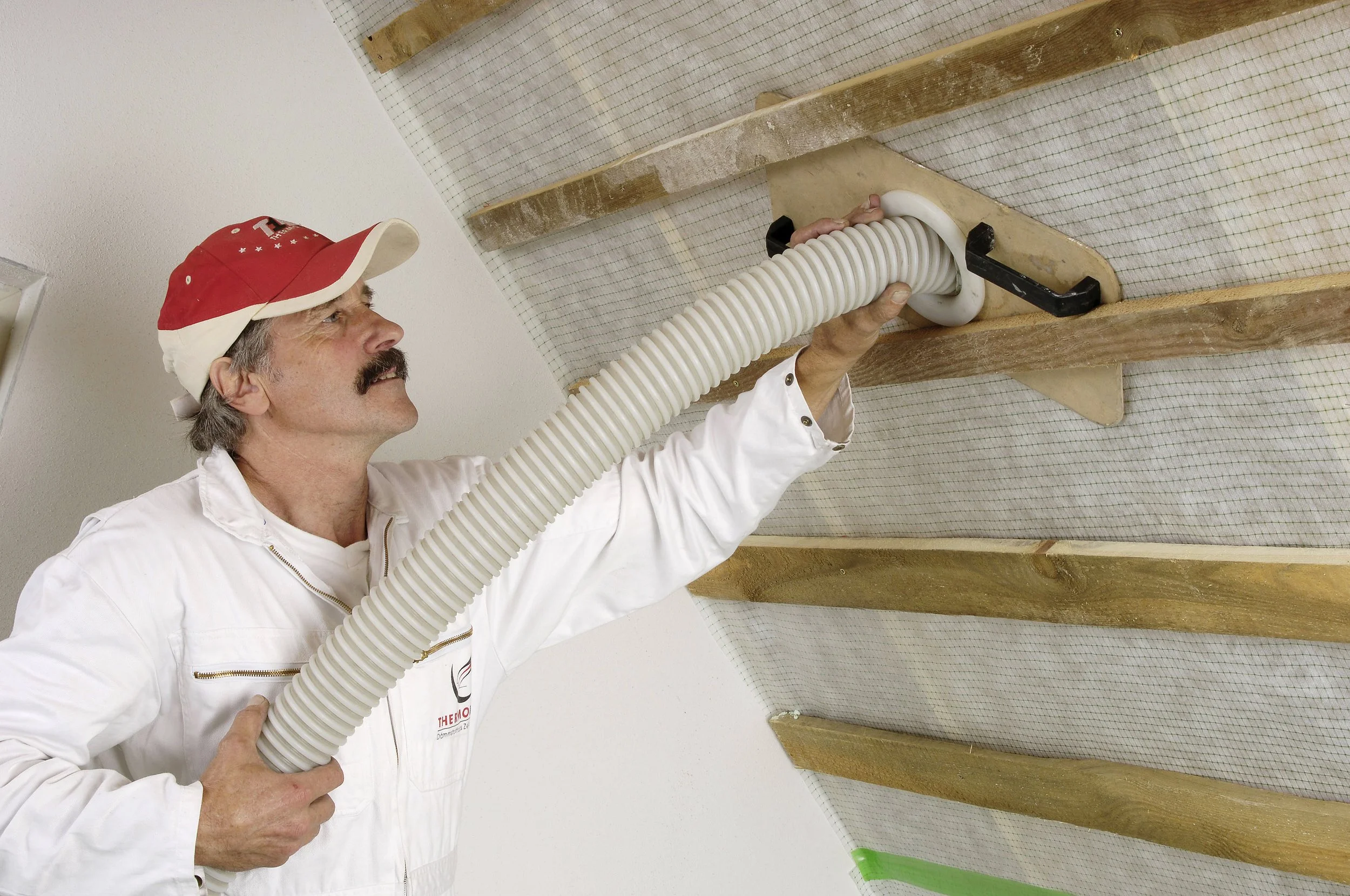 Un homme portant une casquette rouge et un vêtement blanc utilise un tube flexible pour appliquer une substance sur une surface de plafond en bois.
