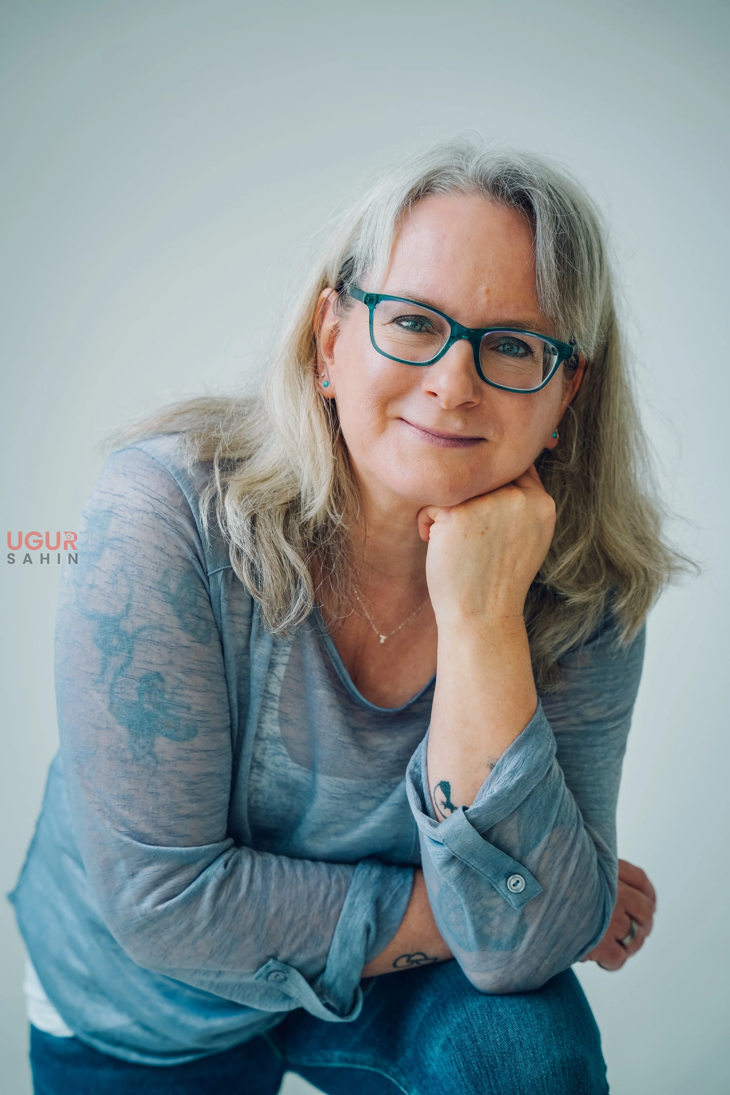 Nahaufnahme einer lächelnden Frau mit Brille, der in die Kamera schaut, auf einem hellen Hintergrund. Sie trägt eine graue Bluse und hat blonde, lockige Haare.