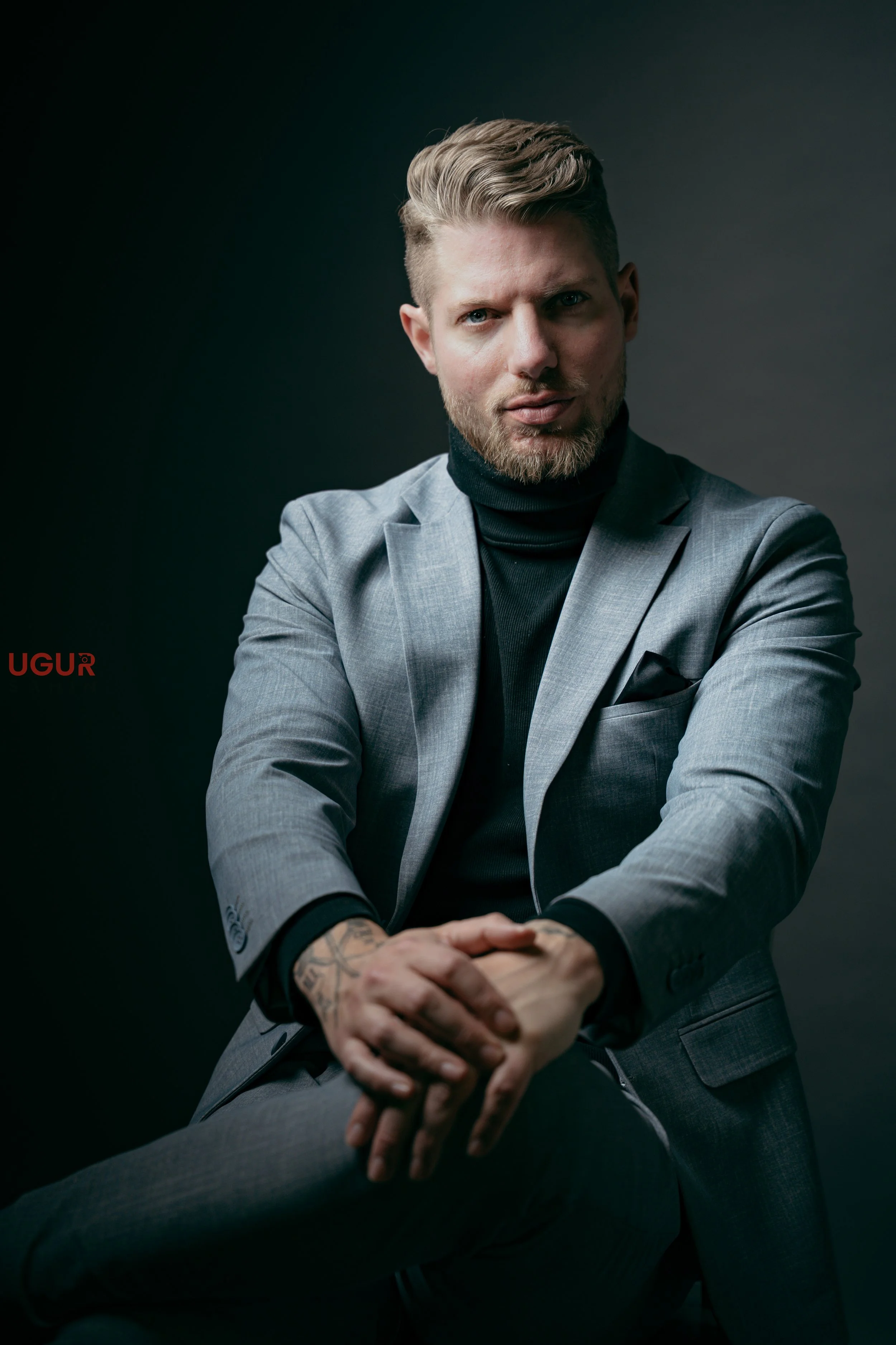 Junger Mann mit blonden Haaren, gepflegtem Bart, dunklem Rollkragenpullover und grauem Anzug, sitzend gegen dunklen Hintergrund, mit erhobenem Arm und verschränkten Händen.