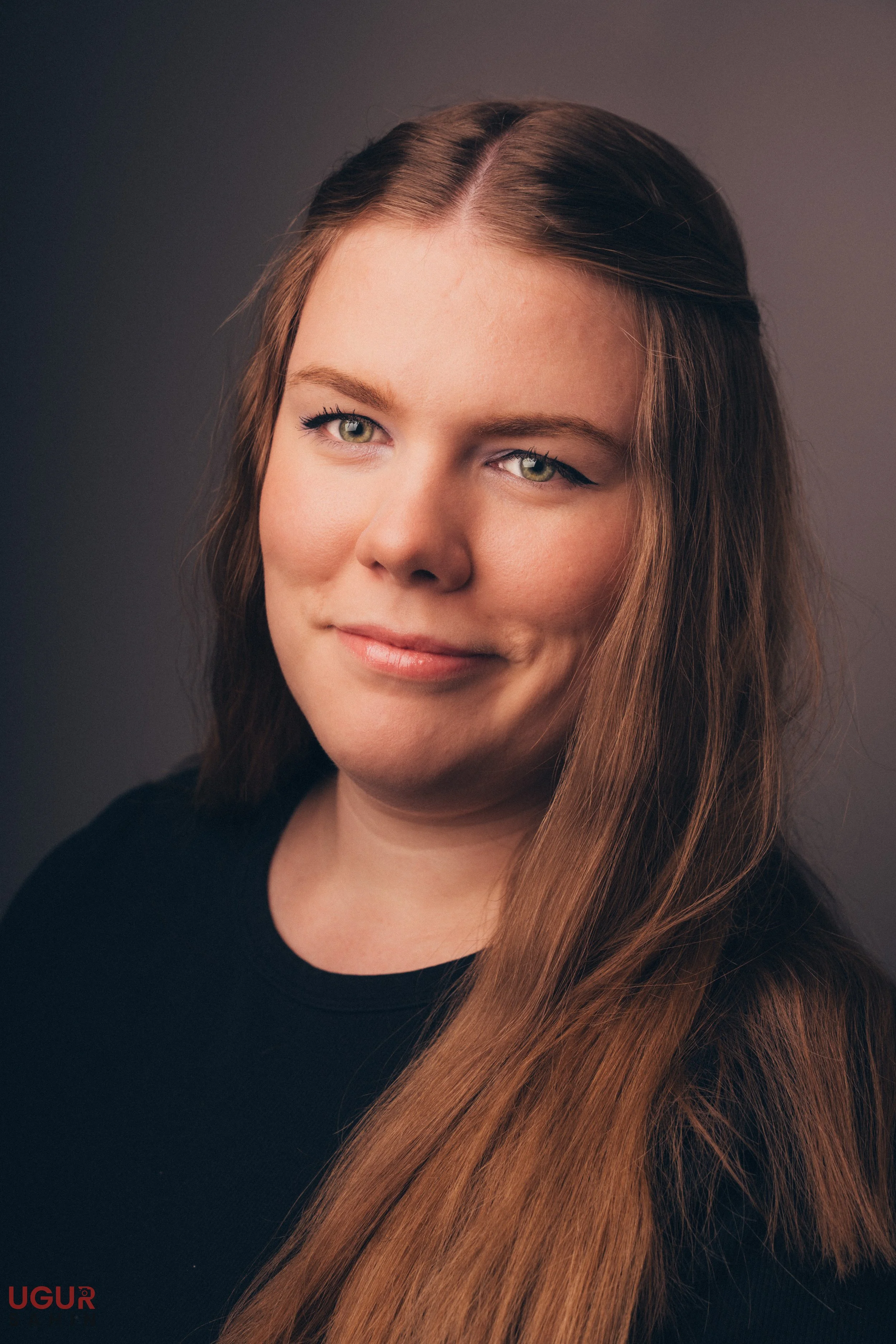 Porträt eines jungen Mädchens mit langen braunen Haaren, die lächelnd in die Kamera schaut, gegen einen grauen Hintergrund.