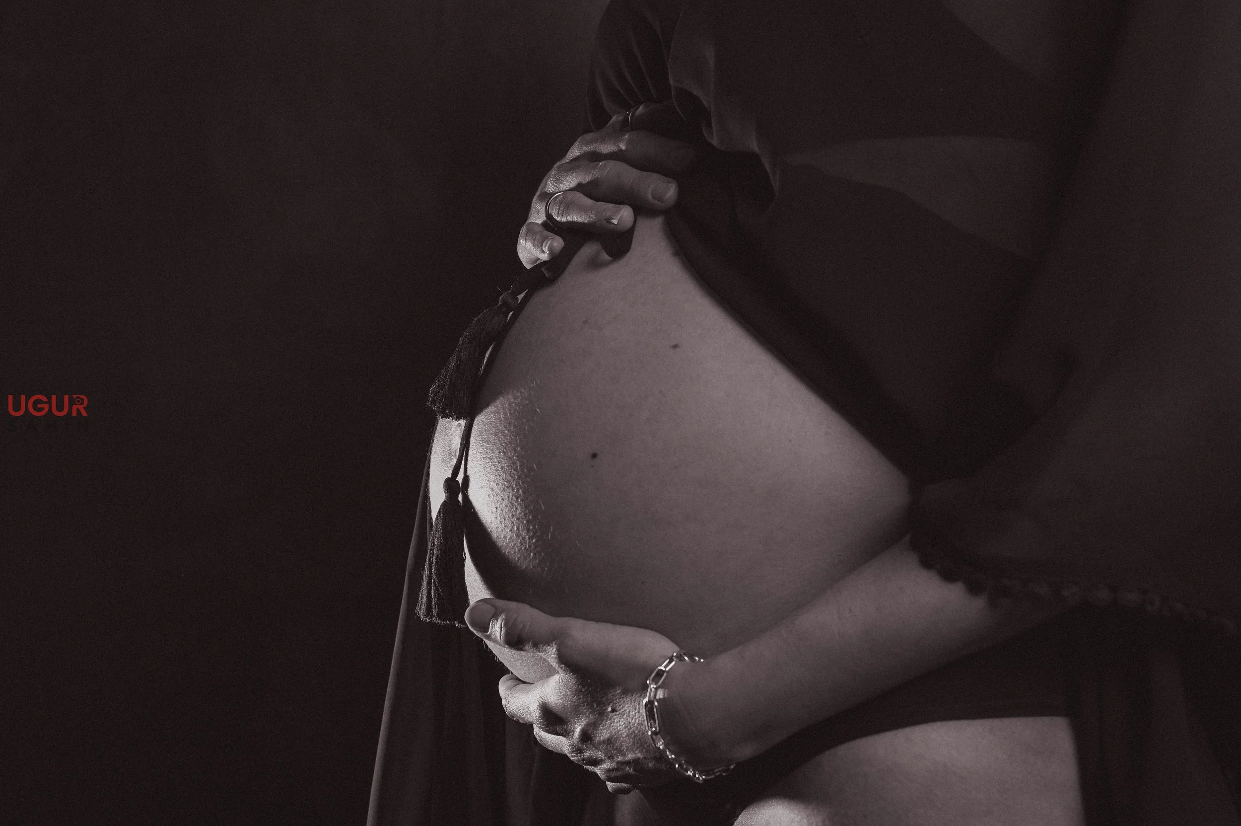Nahaufnahme eines schwangeren Bauches in schwarz-weiß, jemand hält den Bauch mit beiden Händen, es trägt eine Armband und die Person trägt dunkle Kleidung.