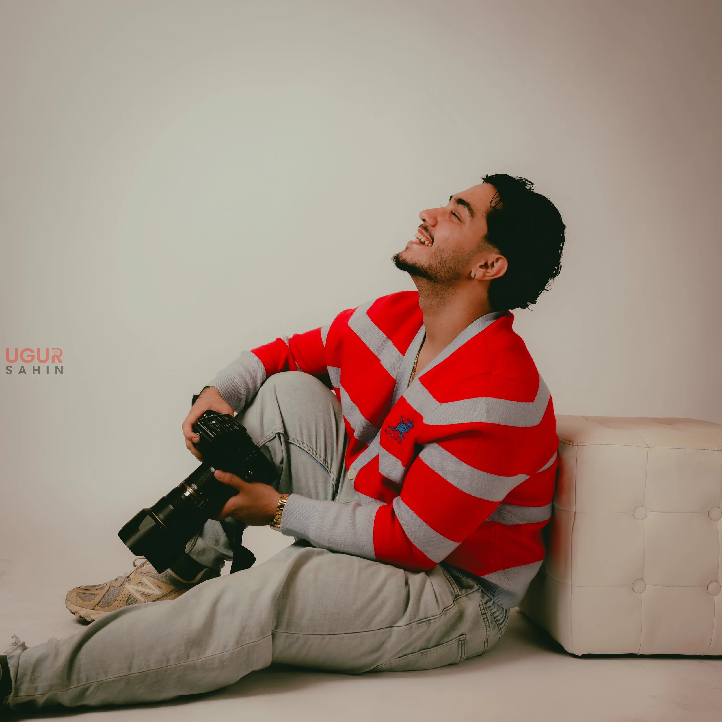 Ein lachender junger Mann sitzt auf dem Boden, trägt eine rote und grau gestreifte Jacke, beige Hosen und hält eine Kamera in der Hand. Hinten ein weißes gepolstertes Möbelstück, der Hintergrund ist schlicht und hell.