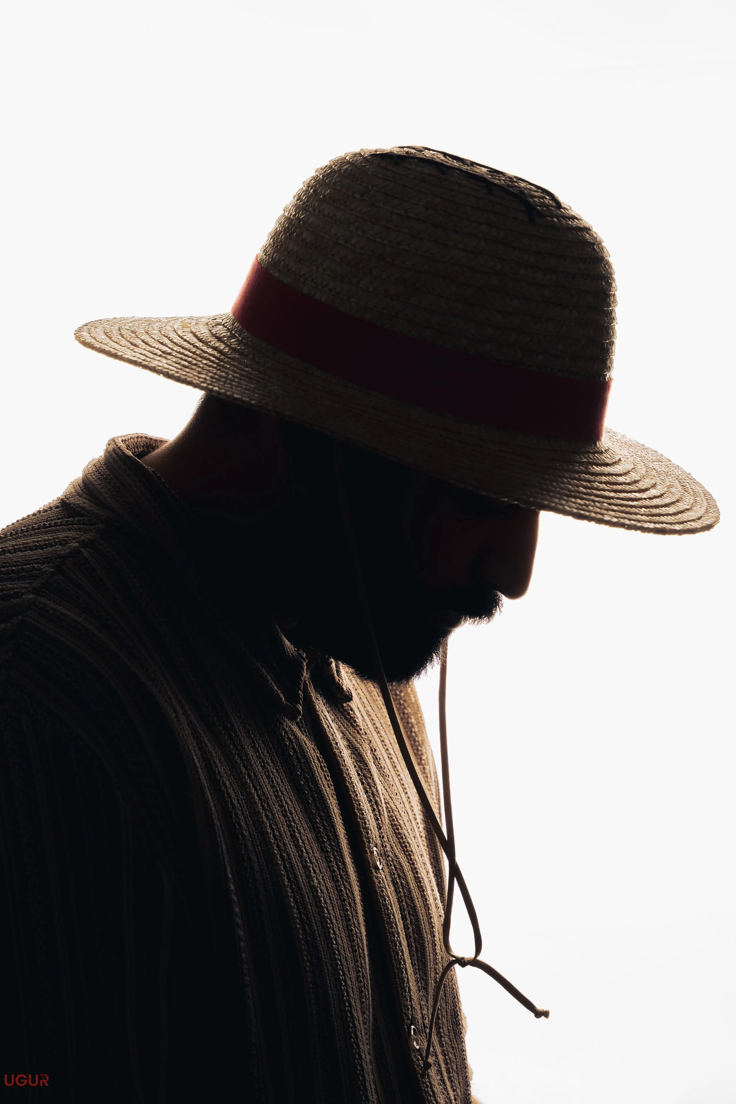 Schattenhafter Porträt eines Mannes mit Strohhut, der nach unten schaut, vor hellem Hintergrund.