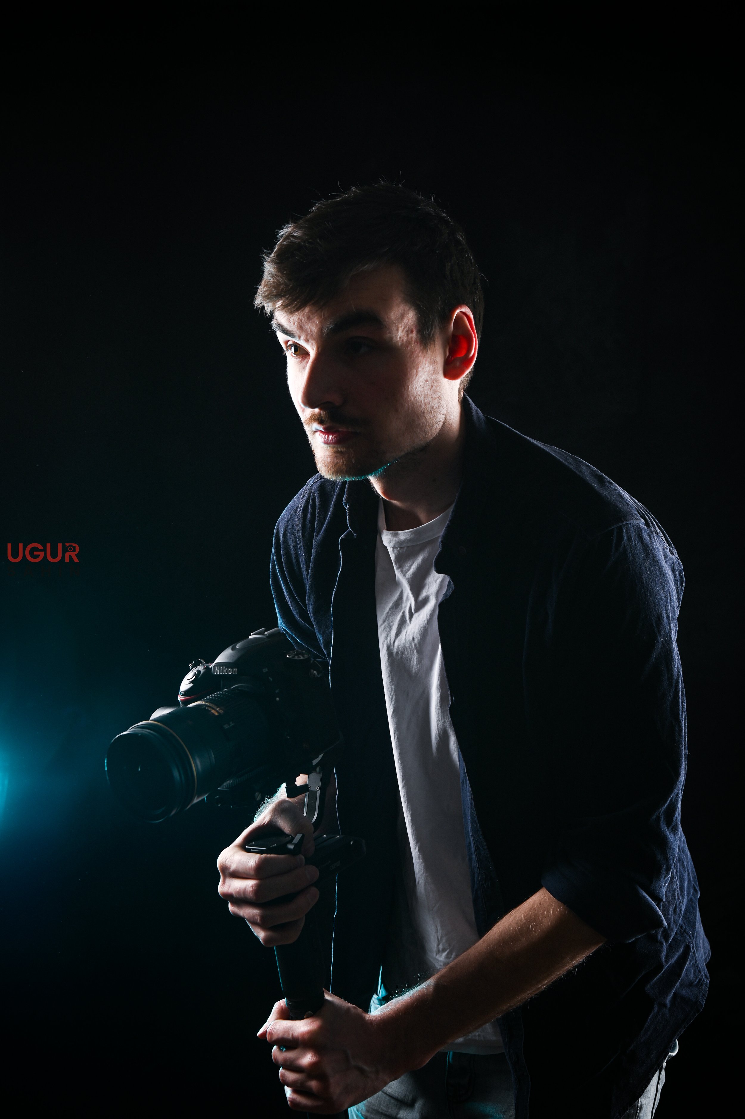 Ein junger Mann hält eine Kamera in der Hand, steht vor einem dunklen Hintergrund, mit seitlicher Beleuchtung, welche sein Gesicht teilweise beleuchtet.