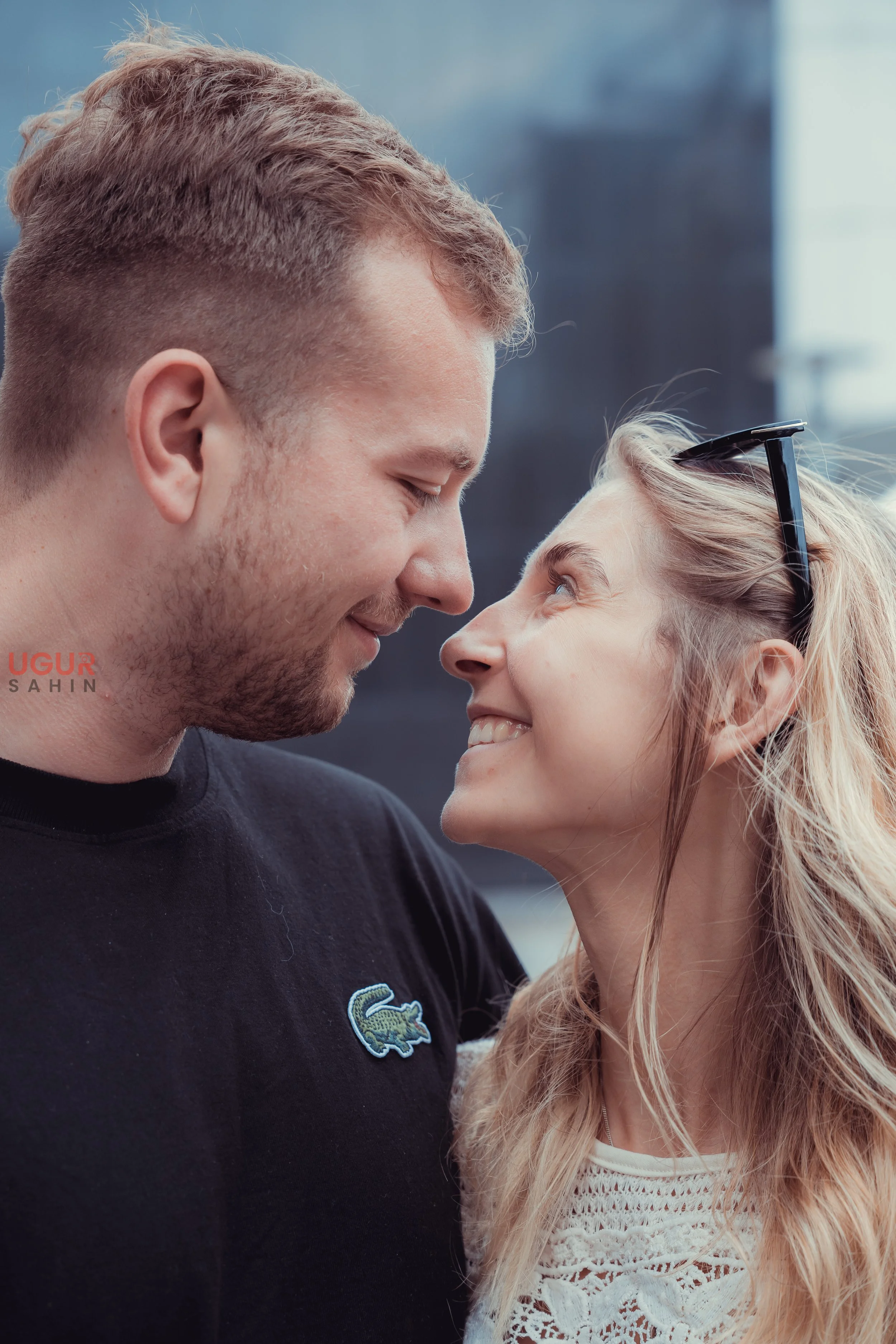 Ein lachendes Paar, das sich sehr nahe steht, schaut sich liebevoll in die Augen, im Freien vor einem unscharfen urbanen Hintergrund.