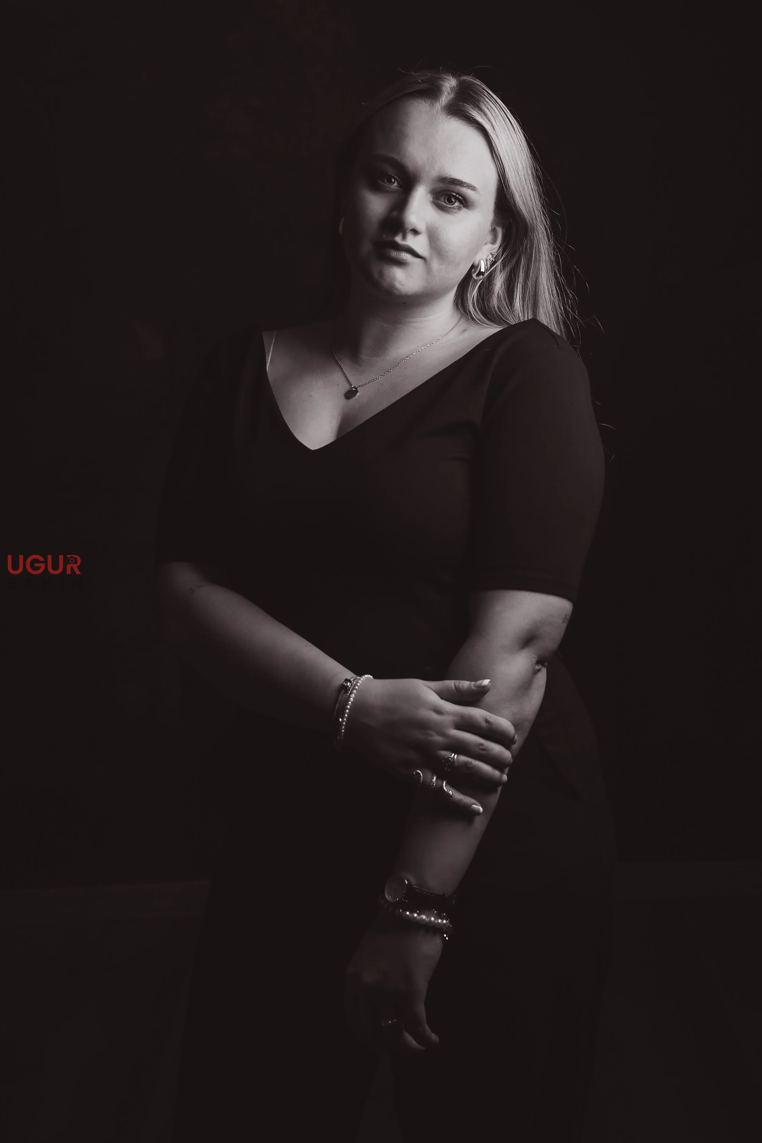 Schwarz-weiß-Fotografie einer jungen Frau mit langen Haaren, die schwarze Kleidung trägt und Schmuck zeigt, vor dunklem Hintergrund.