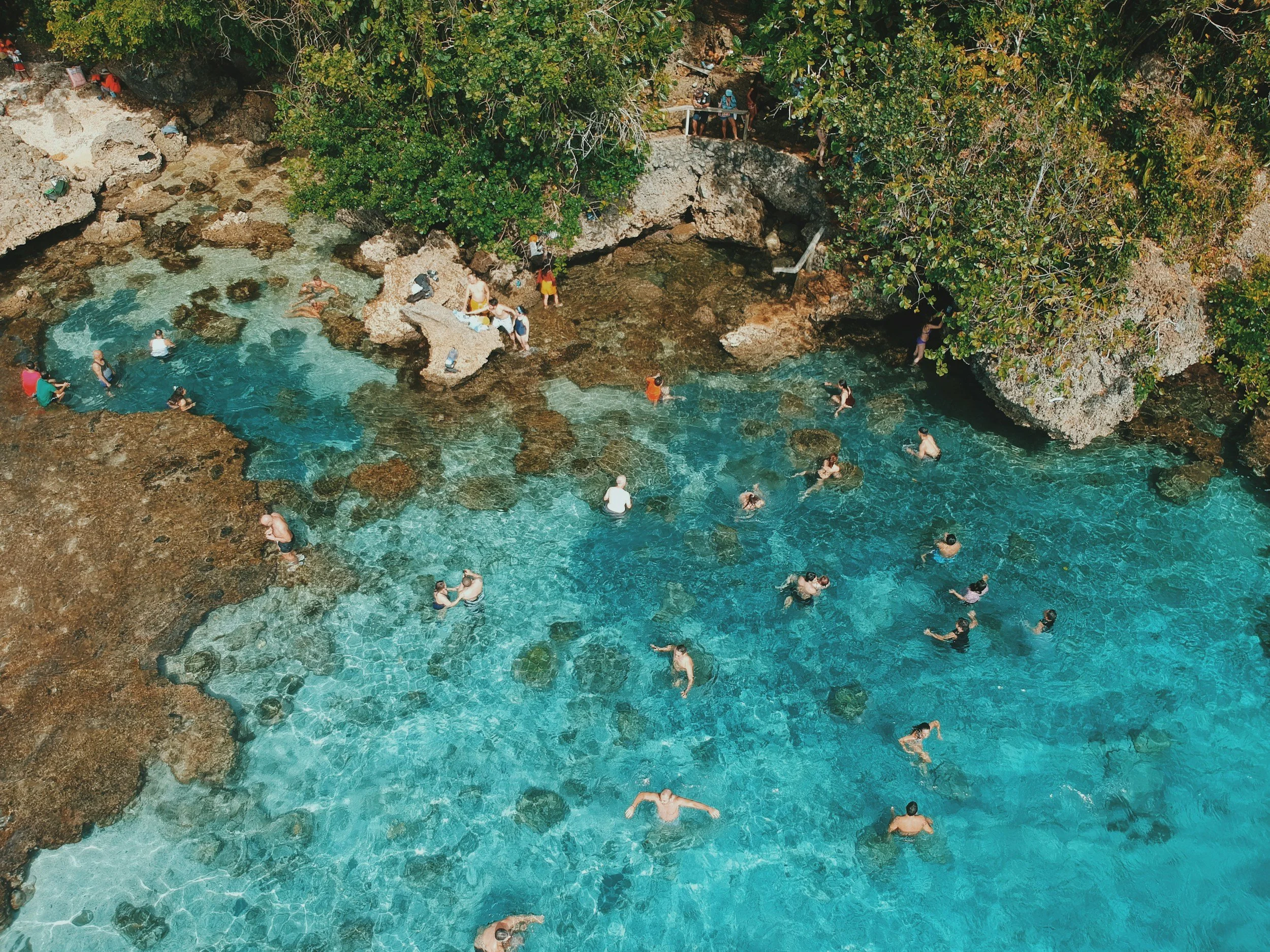 An aerial view of many people swimming and wading in the clear, turquoise water of the Magpupungko Rock Pools in Siargao, surrounded by rugged limestone rocks and green trees.