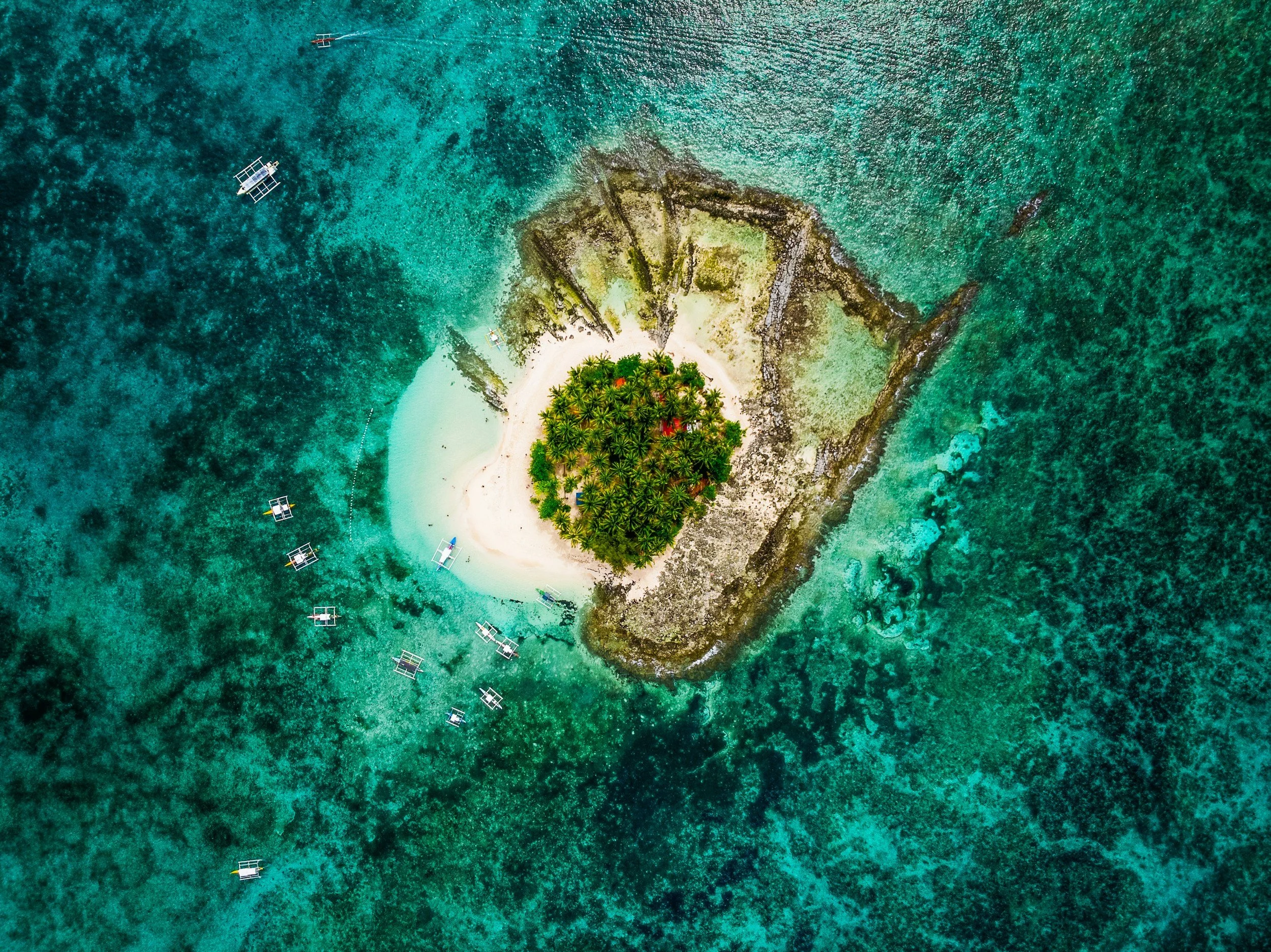 An aerial drone shot of Guyam Island in Siargao showing a small circular patch of green palm trees surrounded by white sand and a vast expanse of turquoise coral reefs with several small boats anchored nearby.