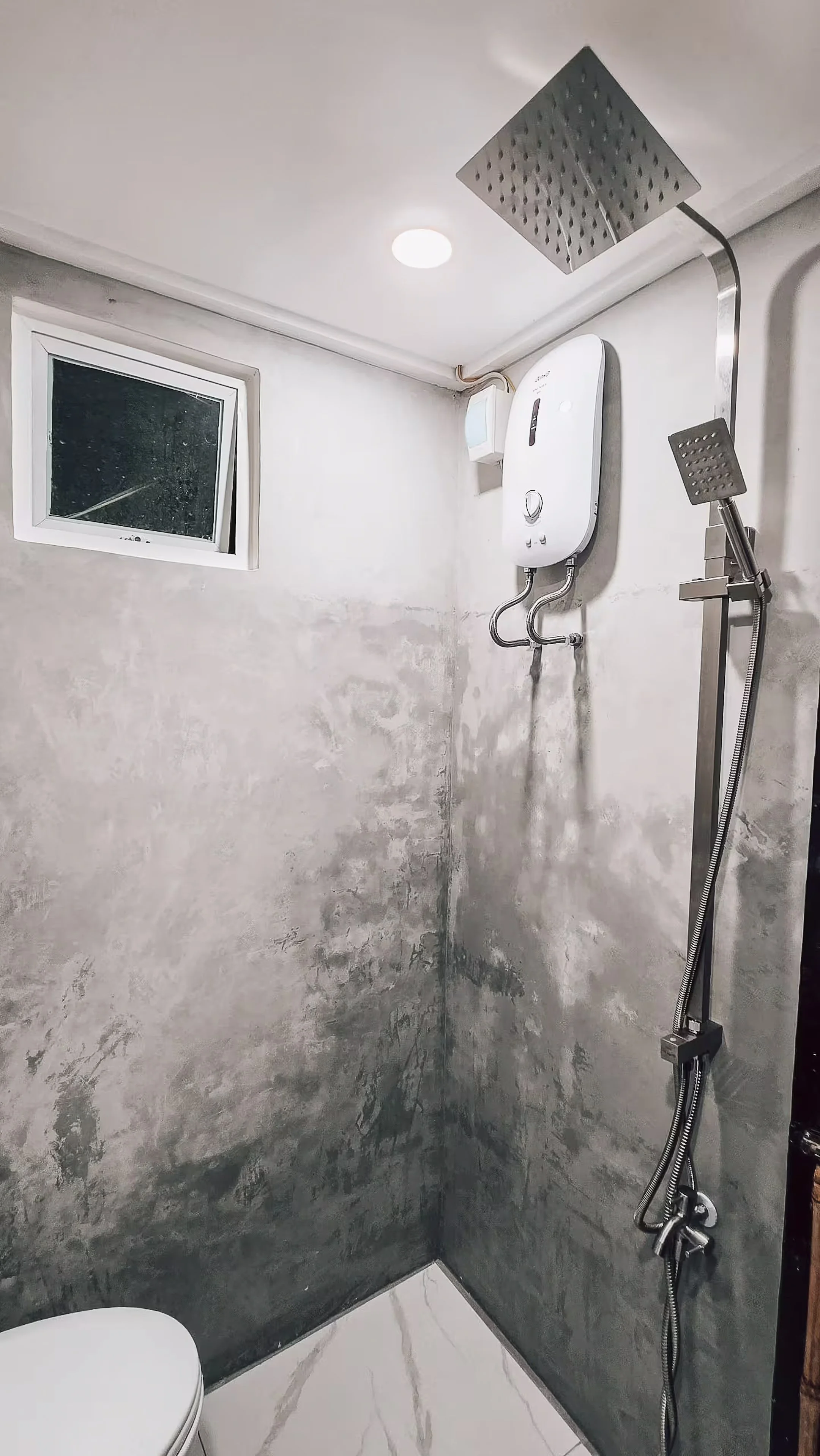 The private bathroom of Surf & Chill Room 3 features a rainfall shower, water heater, and polished concrete walls, offering guests a clean and comfortable space to unwind after exploring Siargao.