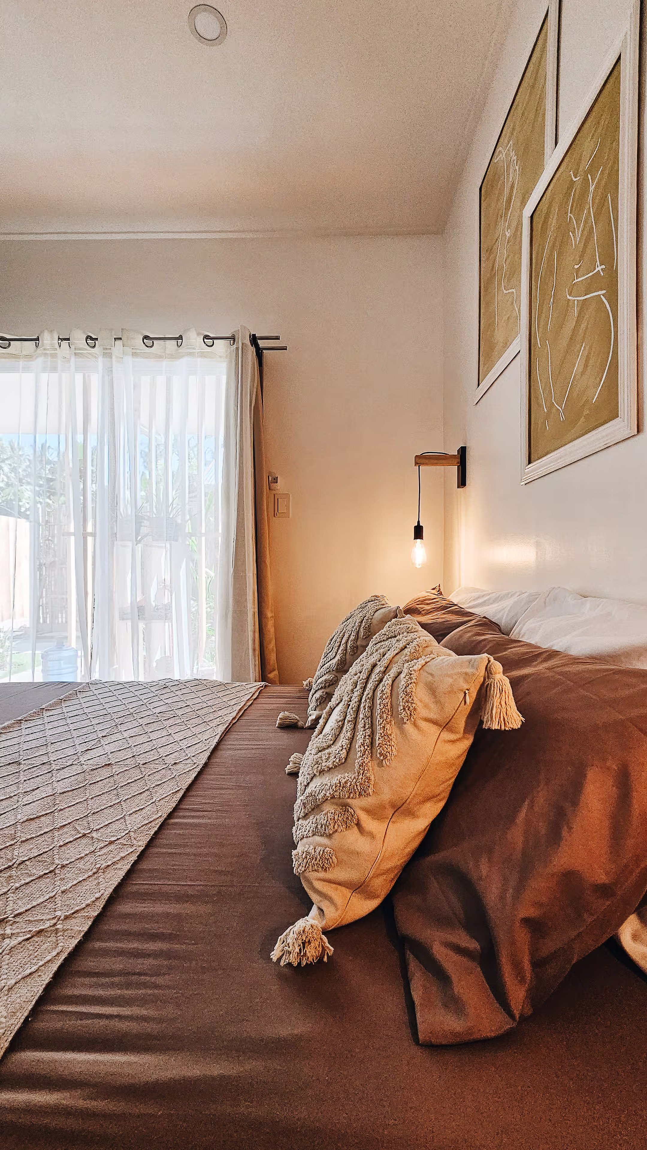 Soft light filters through the curtains of Room 1, revealing a calm space with a queen bed, textured pillows, and earth-toned bedding. Perfect for slow mornings and quiet nights in General Luna.