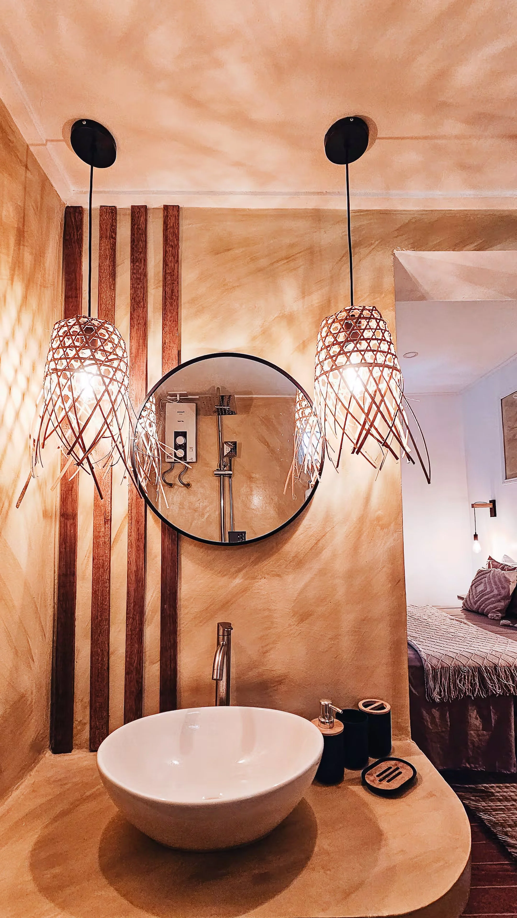 Soft light spills from woven rattan lamps over a minimalist vanity with a ceramic basin and round mirror. A warm, island-inspired touch that connects the bedroom and bath in Room 1.