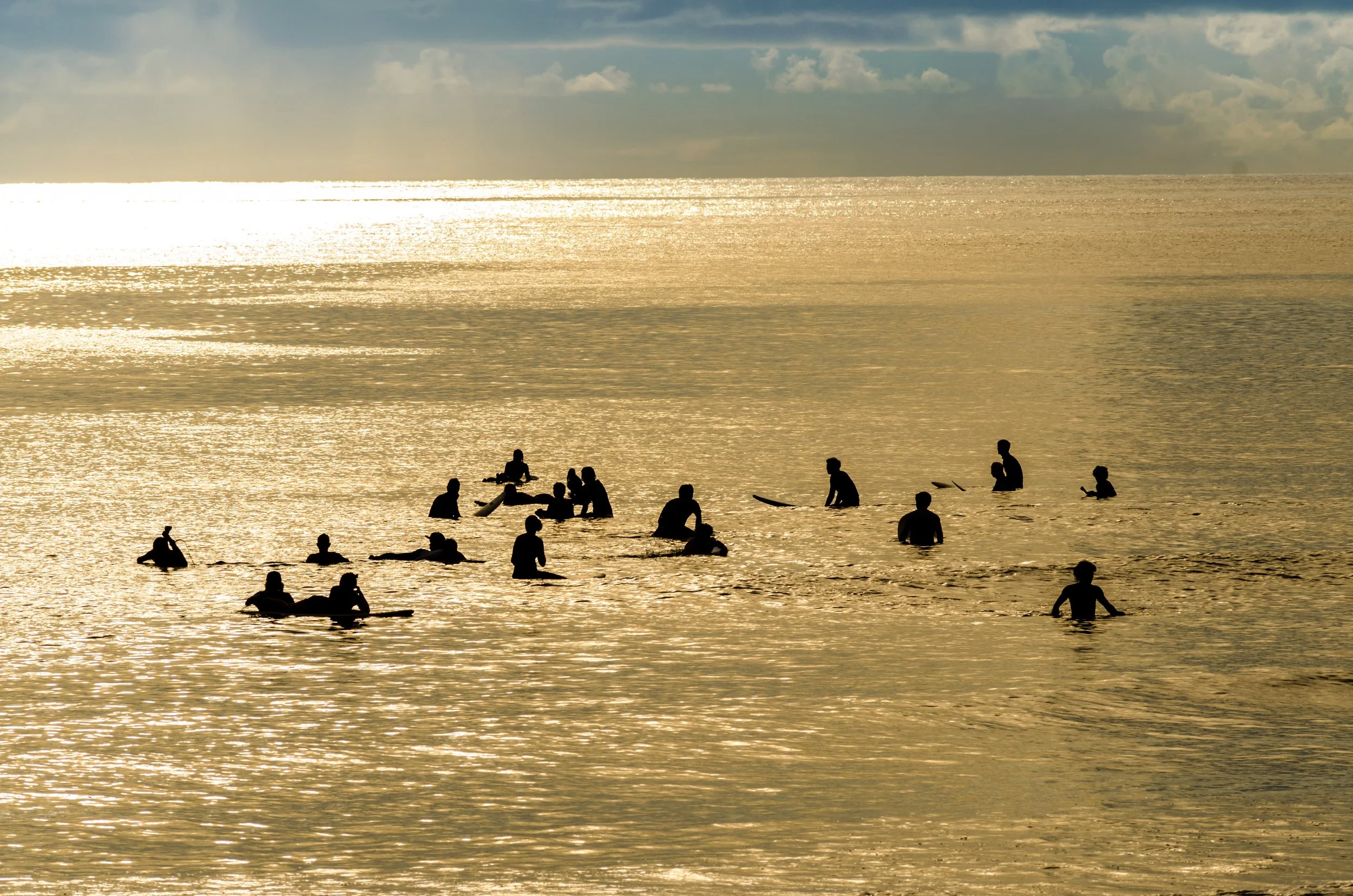 Silhouettes of several surfers sitting on their boards in the ocean at Siargao during a golden sunset with bright sunlight reflecting off the water.